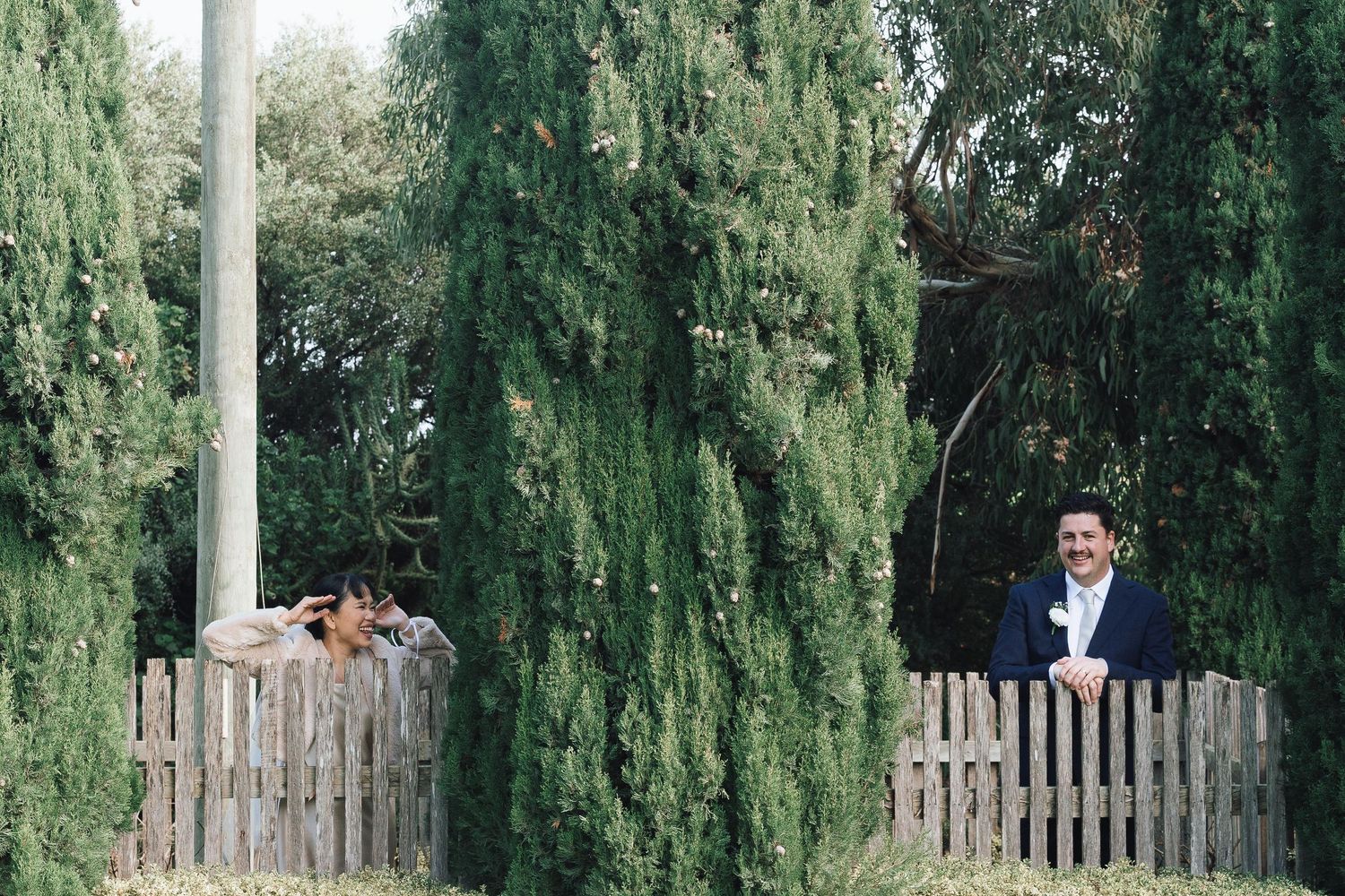 Groom in a navy suit standing behind a wooden picket fence surrounded by tall cypress trees.