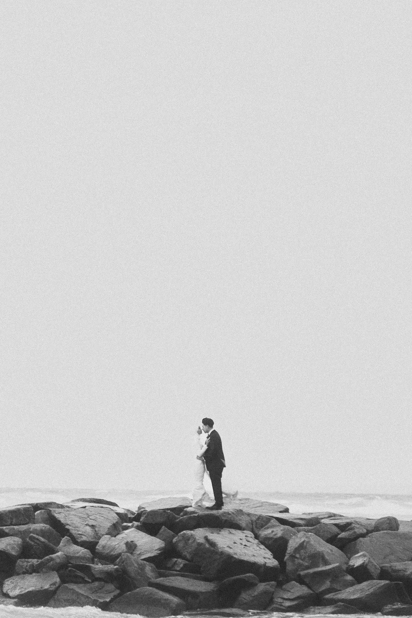 Black and white photo of bride and groom standing on rocky shoreline embracing against overcast sky.