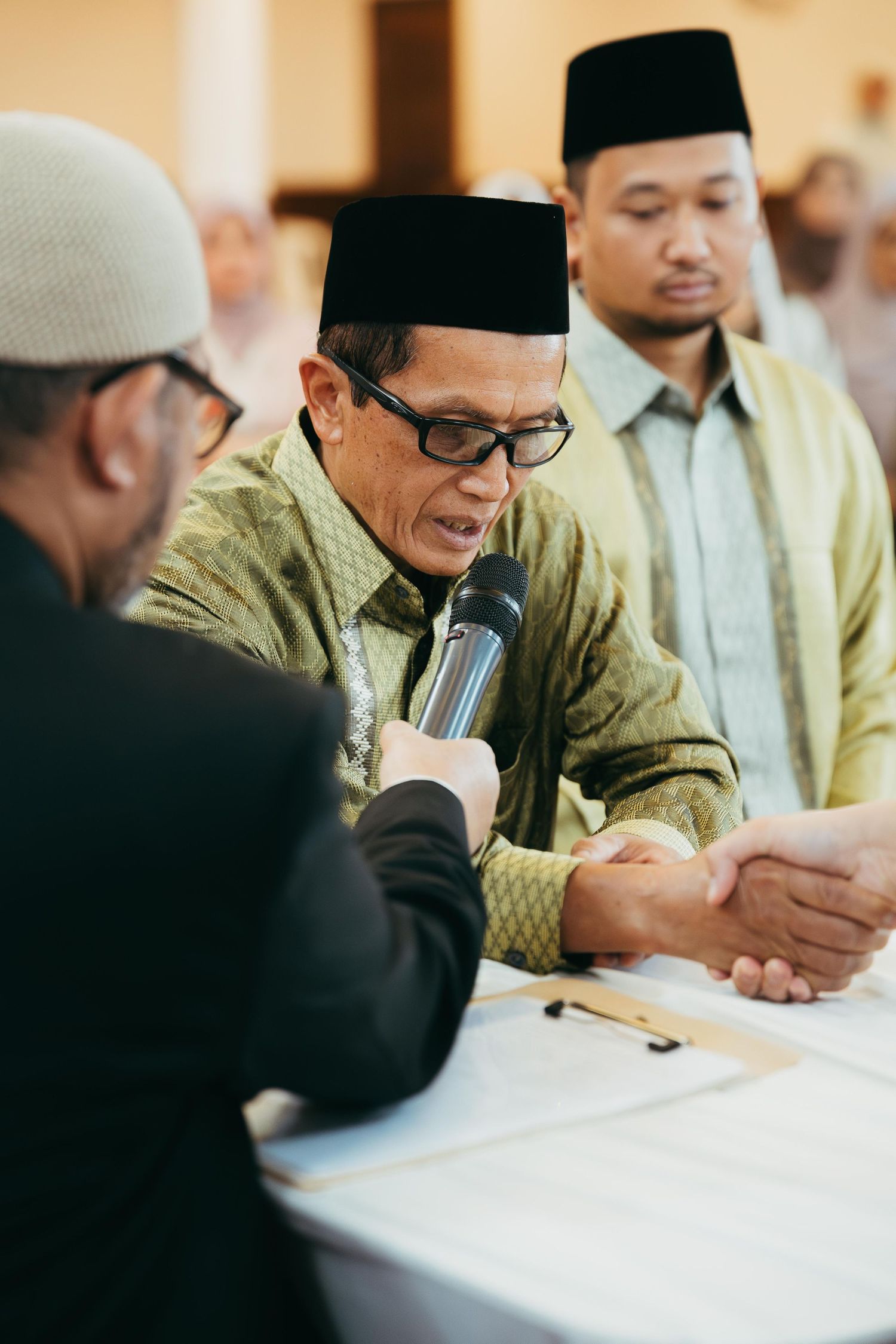 Elder Muslim man in traditional cap speaks into microphone while shaking hands during Islamic wedding Nikah ceremony.