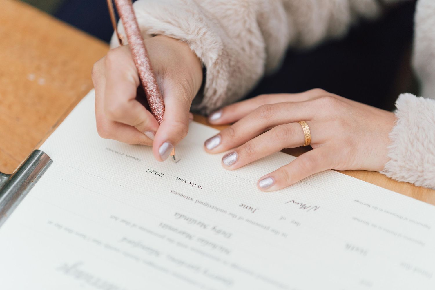 Close-up of a woman signing a document with a pink pen, wearing a gold ring and neutral manicure.