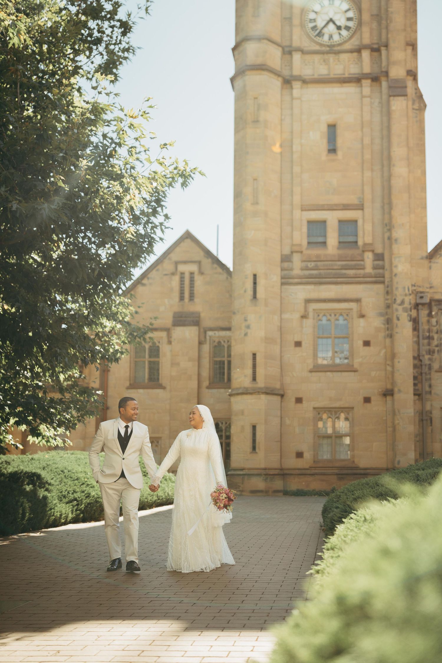 Bride in white hijab gown and groom in beige suit holding hands before historic stone clock tower.