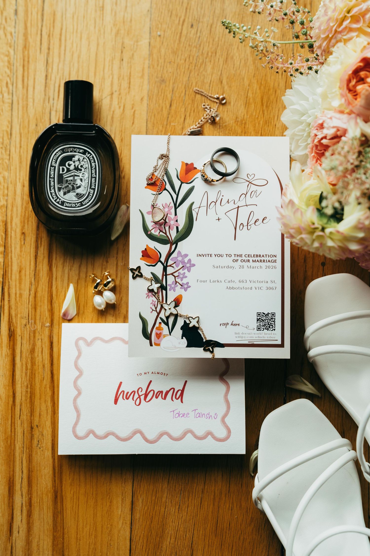 Wedding flat lay with floral invitation, rings, pearl earrings, white sandals, perfume bottle, and pastel bouquet on wood.