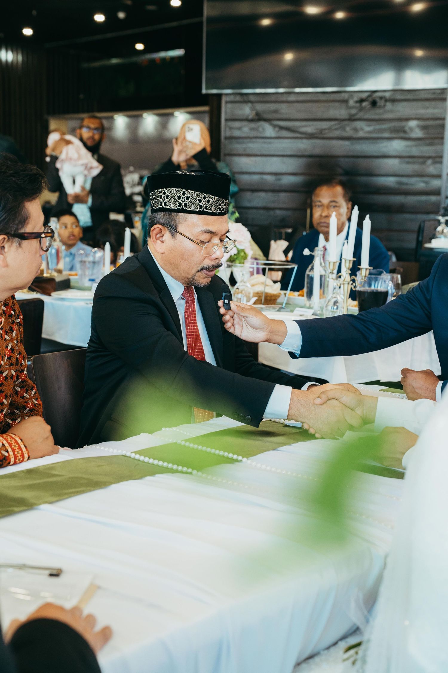 Man in traditional black cap and suit shaking hands at formal wedding reception dinner table.