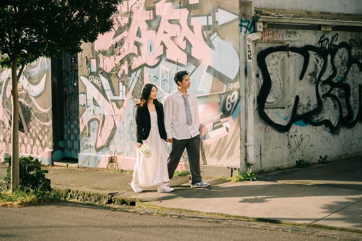 Couple walking and smiling at each other in front of a colorful graffiti wall on a sunny day.