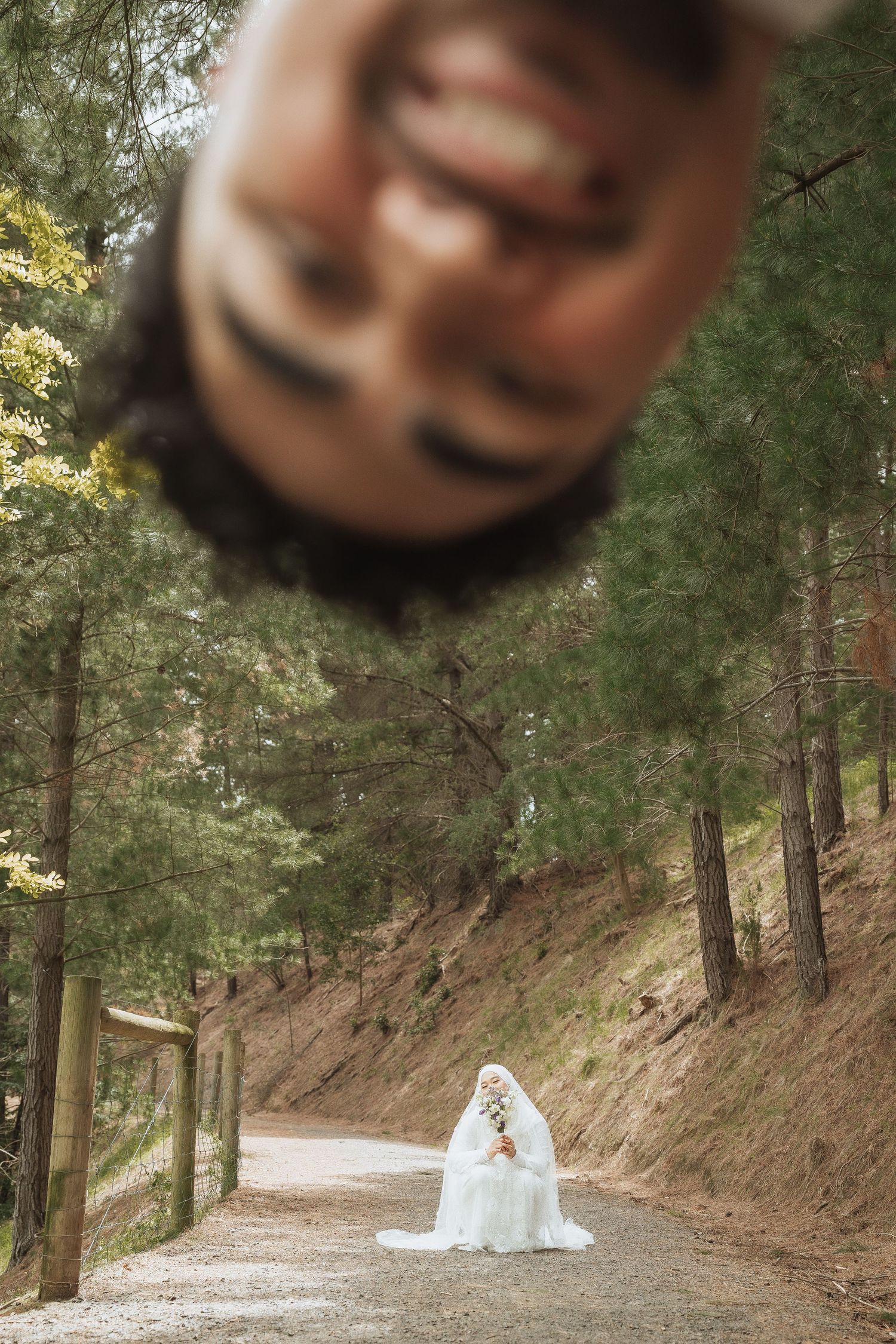 Groom upside down in foreground with bride in white dress sitting on forest path surrounded by tall pine trees.