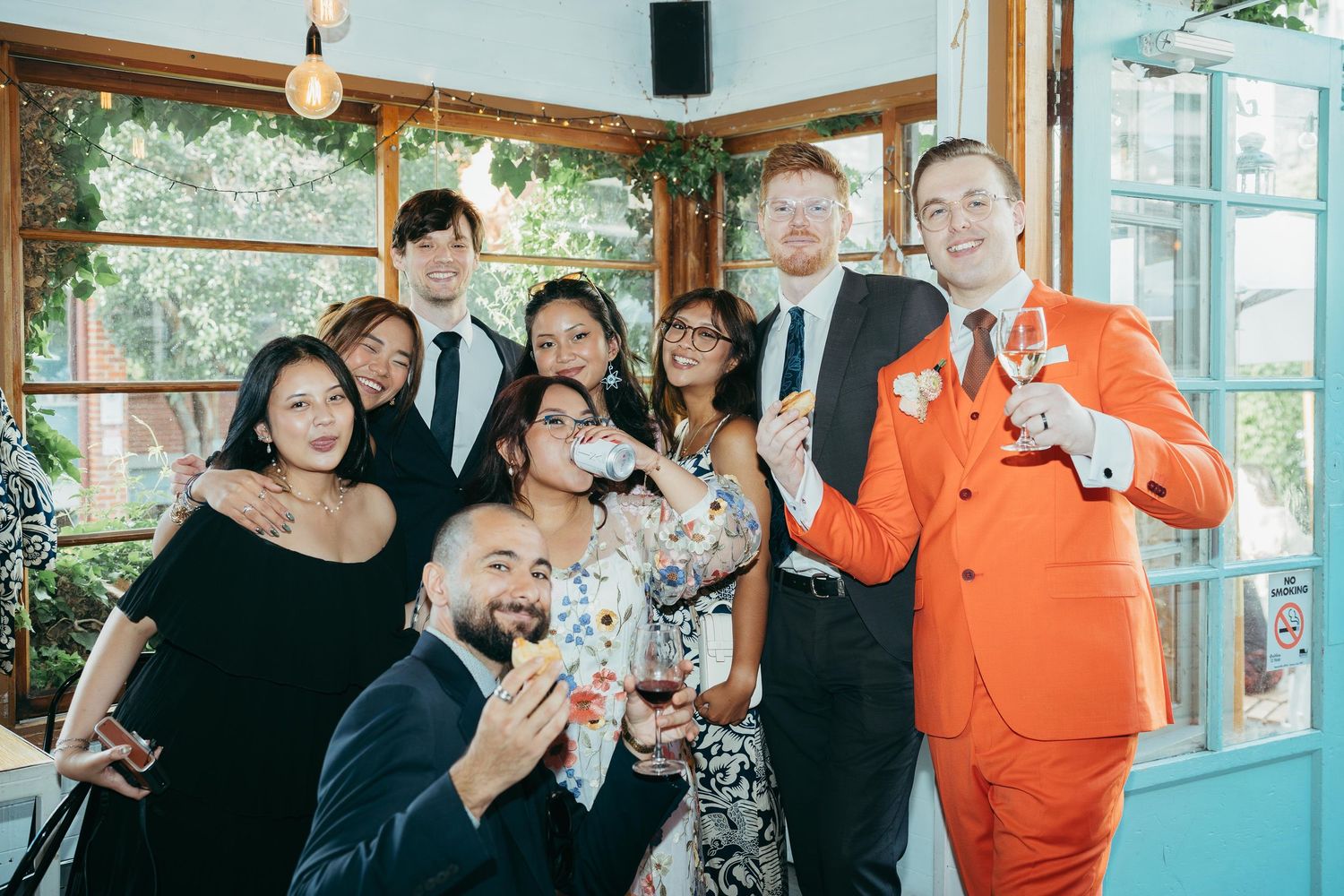 Wedding group photo with groom in orange suit and guests smiling inside a bright reception venue.