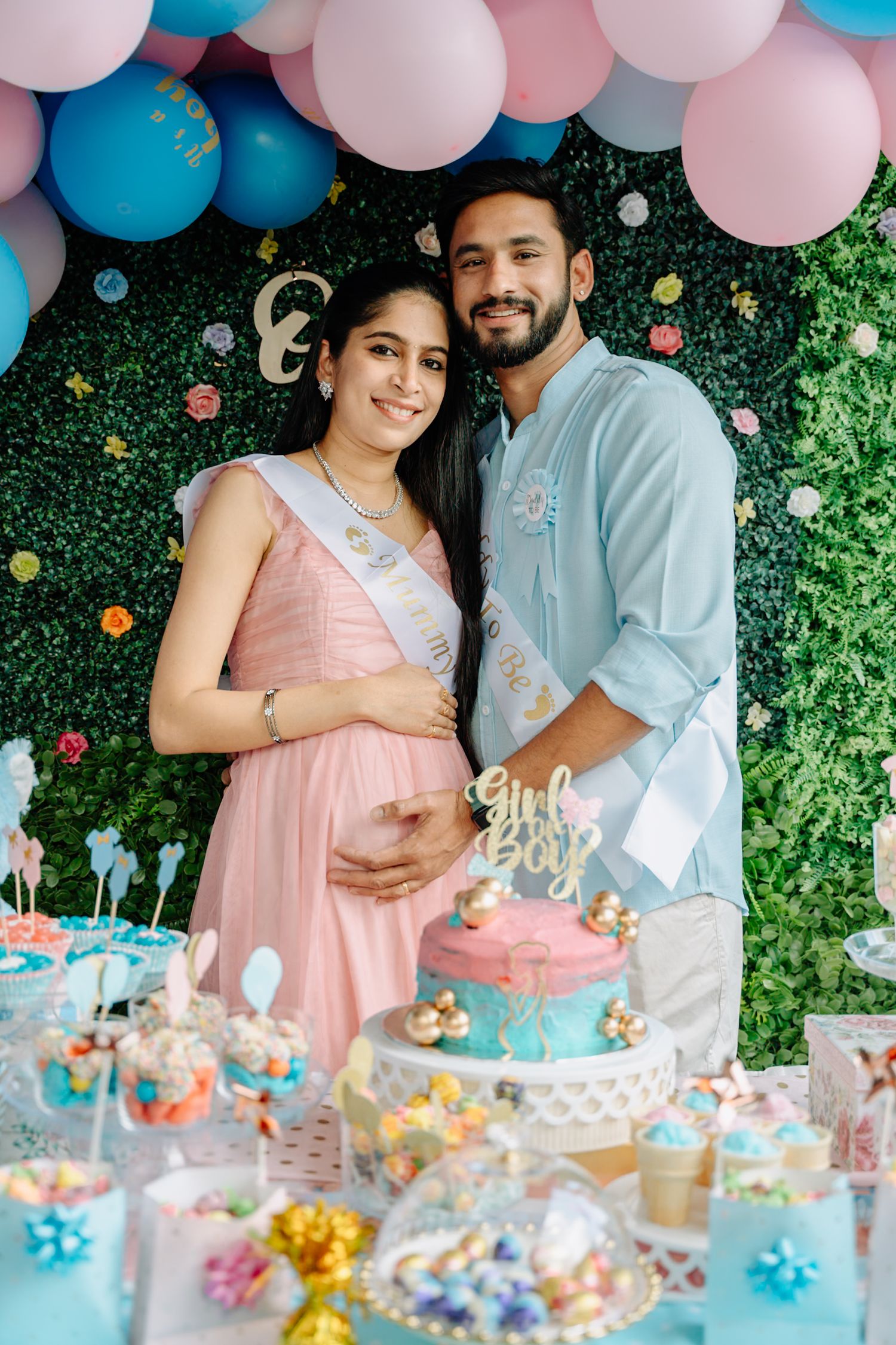 Gender reveal party setup with pink and blue balloons above a dessert table with treats.