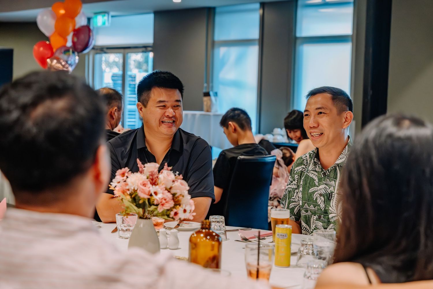 A group gathers around a table decorated with orange balloons for a celebratory meal in a bright modern space.