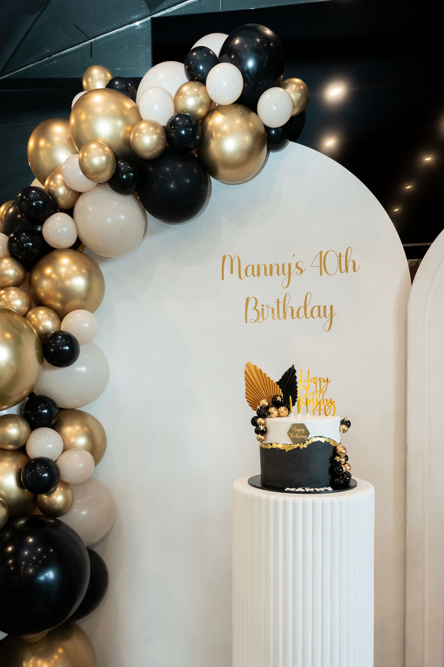 Elegant 40th birthday party setup with black and gold balloon arch and decorative cake on white pedestal.