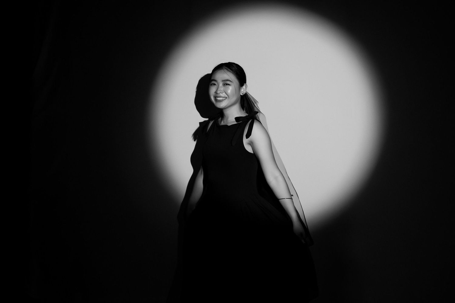 Black and white portrait of a smiling woman in a black dress posed against a dramatic spotlight background.