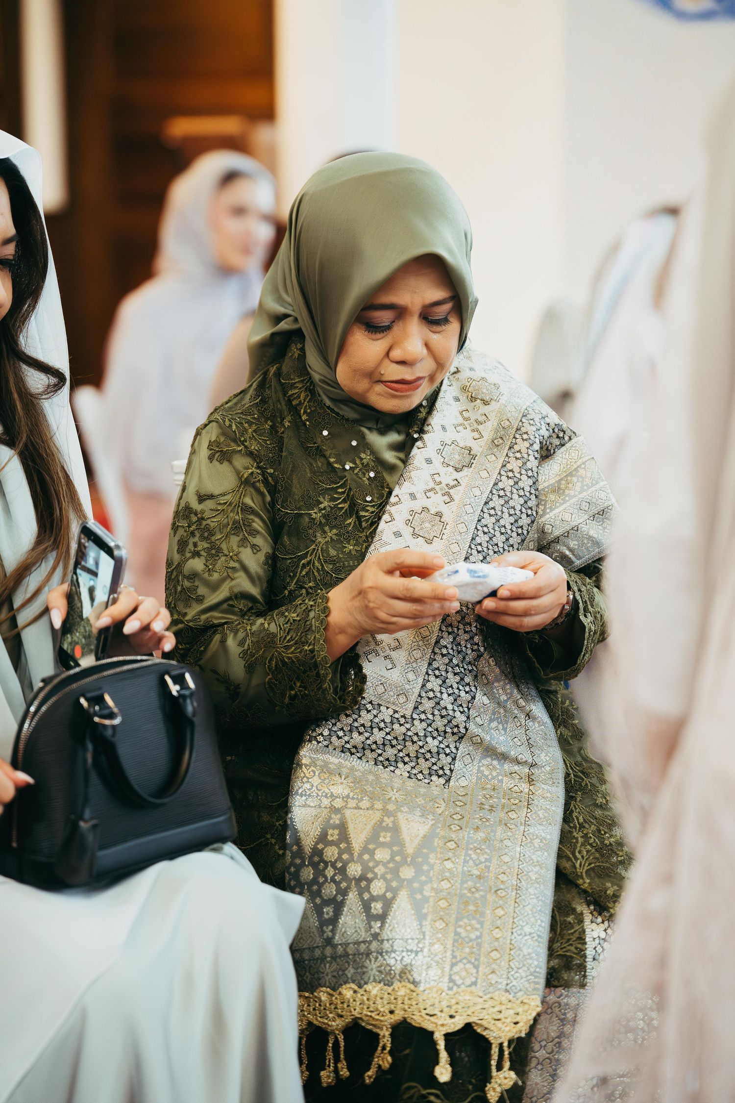 Elderly Muslim woman in green hijab and embroidered outfit examining item at a wedding celebration.