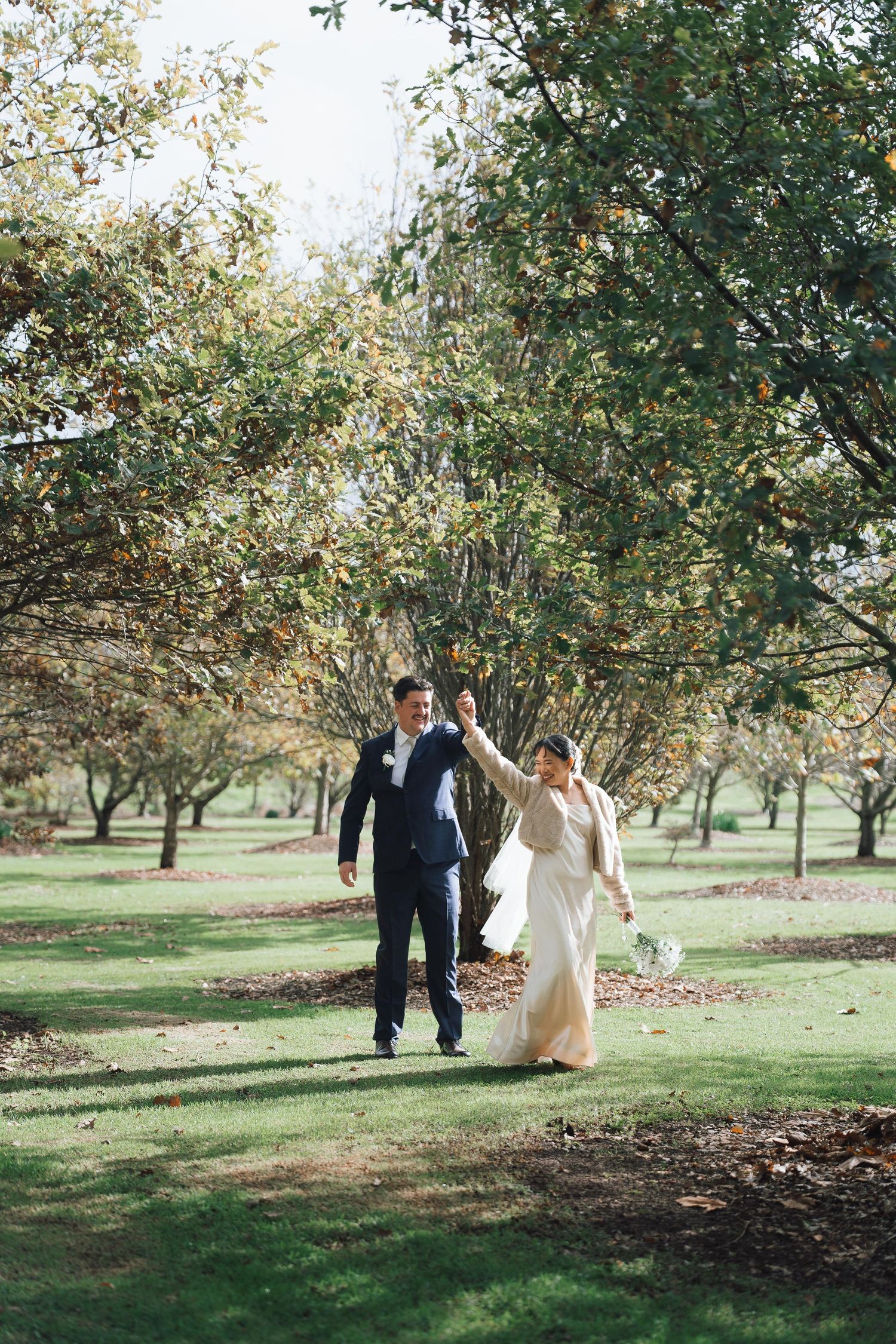 Bride and groom walk hand in hand through a lush green orchard on their wedding day.