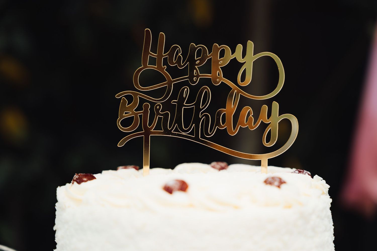 White birthday cake with gold 'Happy Birthday' topper and pink roses on a black background.