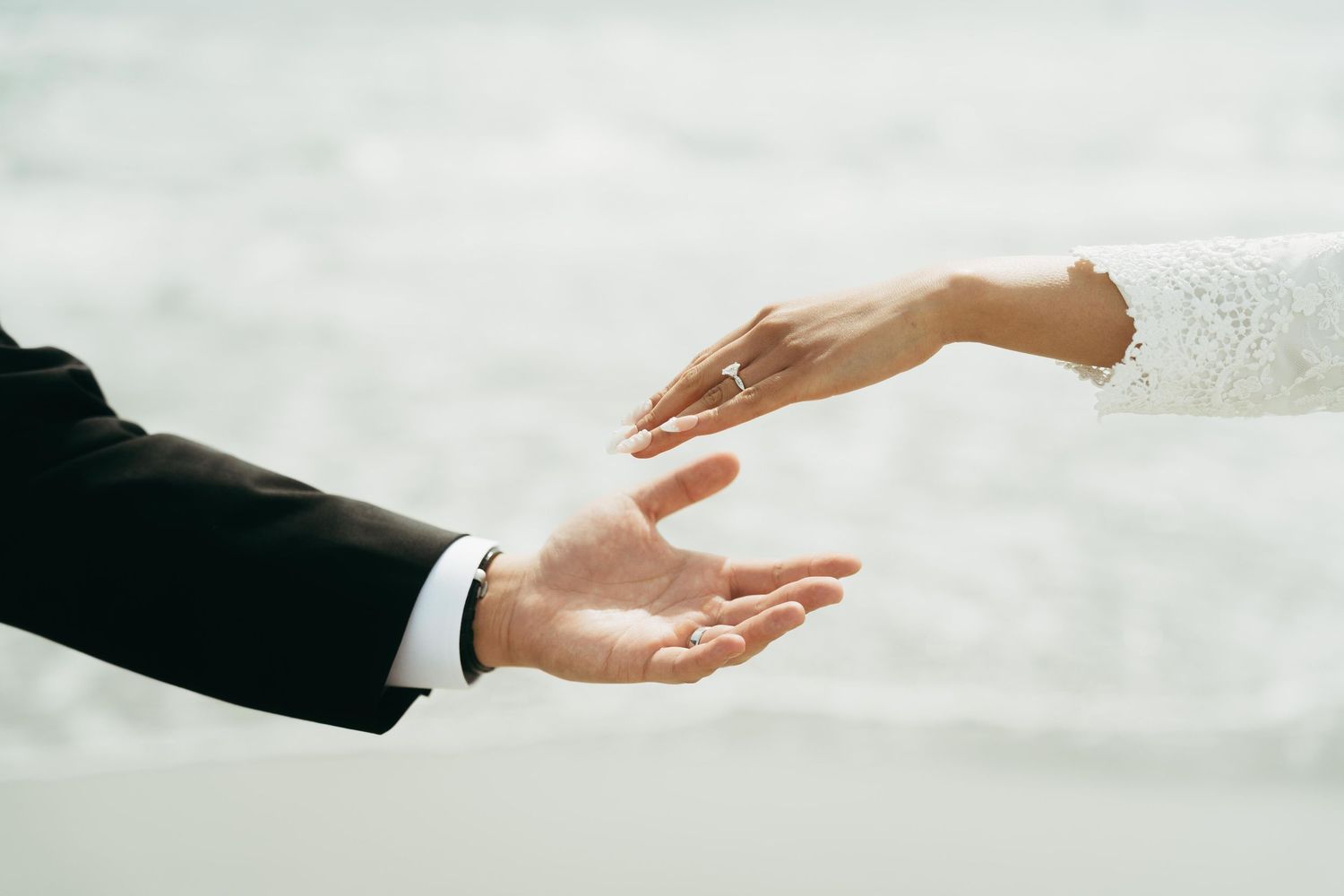 Bride and groom reach for each other's hands near the ocean on their wedding day.