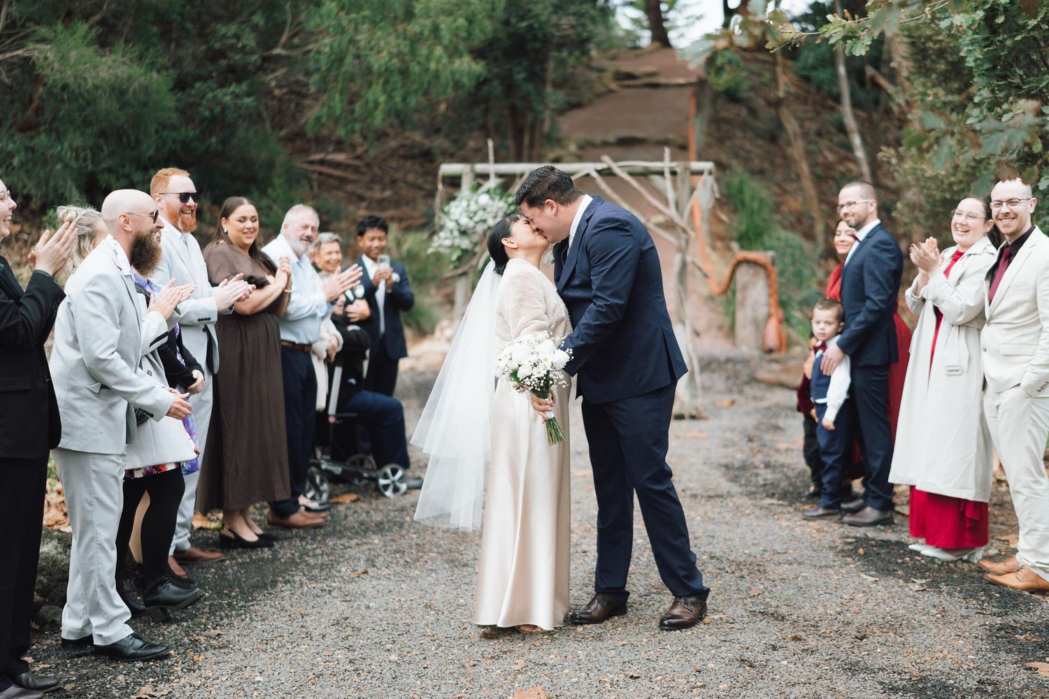 Bride and groom share a kiss while guests celebrate their wedding ceremony exit on a gravel path.