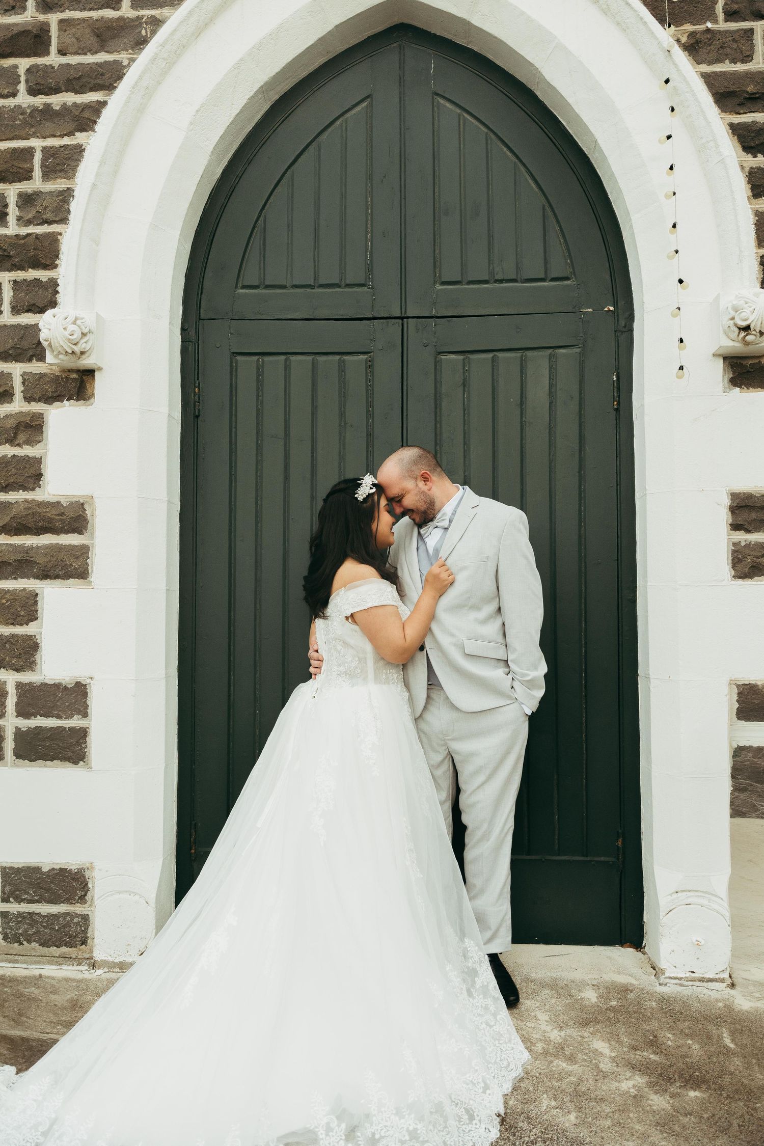 Bride and groom share a romantic kiss in front of a grand arched green church door on their wedding day.