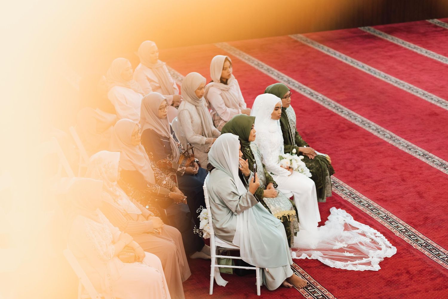Muslim women in hijabs seated at a wedding ceremony inside a mosque with red carpet.