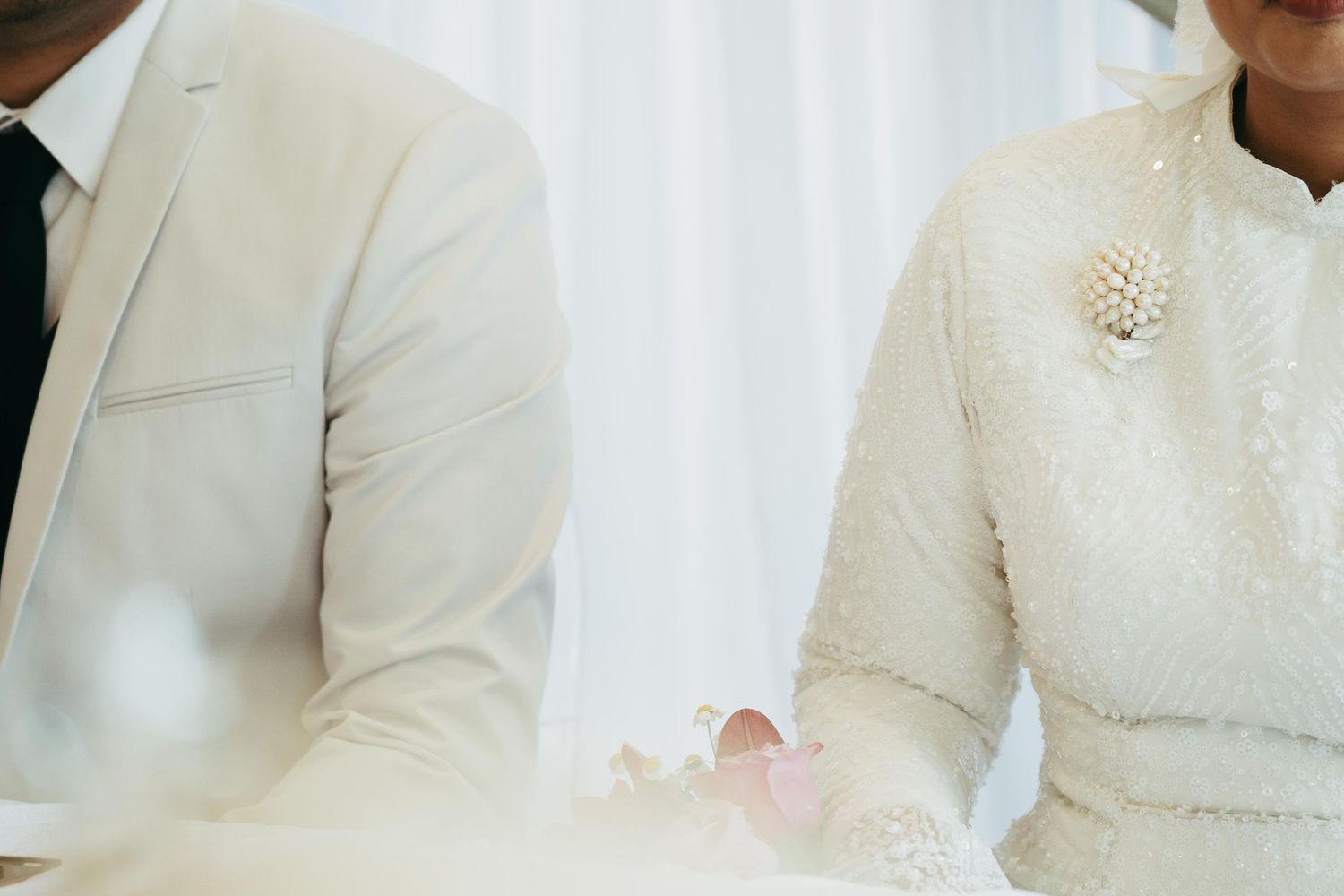 A couple in elegant white wedding attire holding hands, with the bride wearing a pearl brooch at a formal ceremony.
