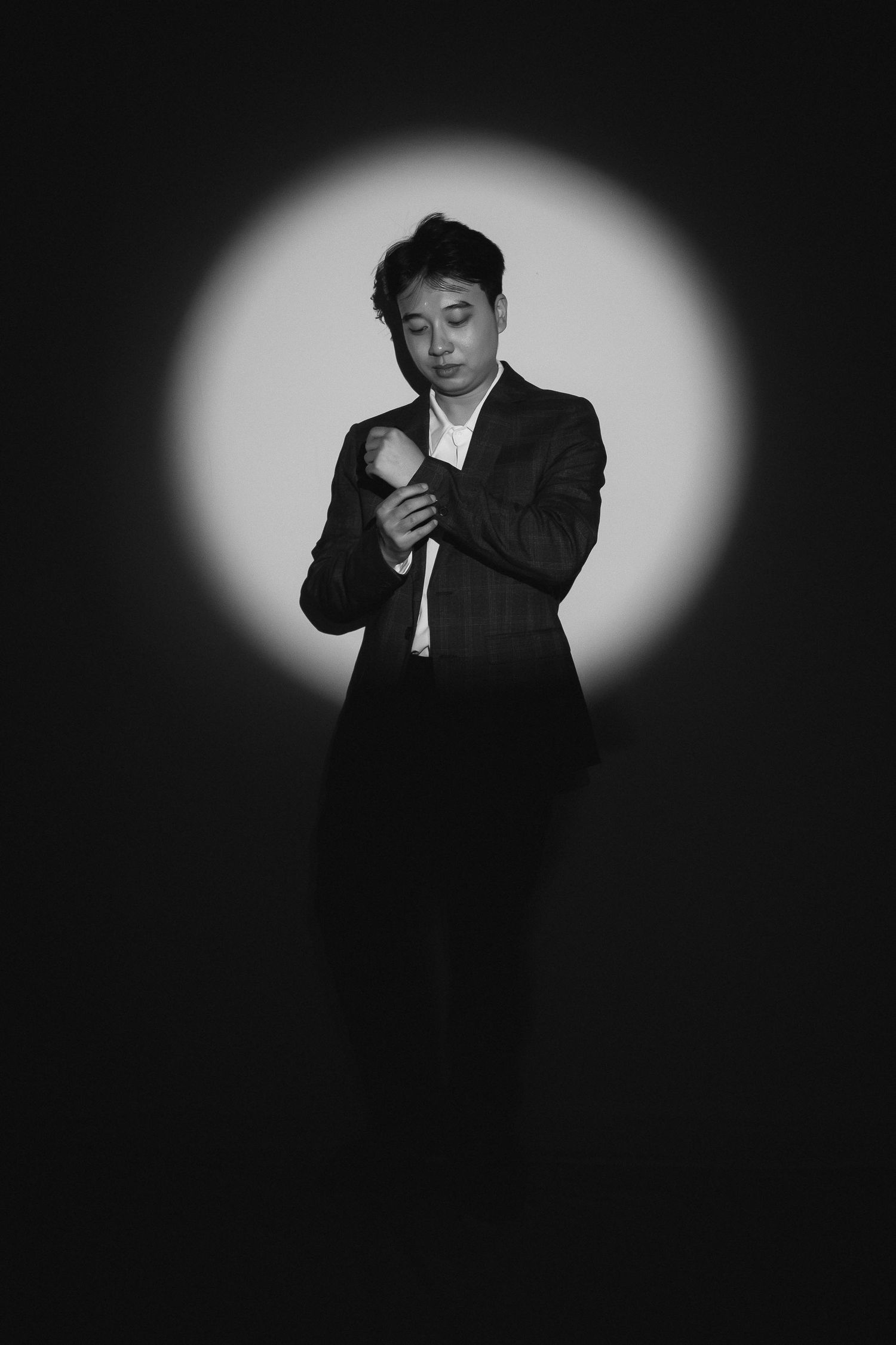 Black and white portrait of a man in a suit adjusting his sleeve under a circular spotlight on dark background.