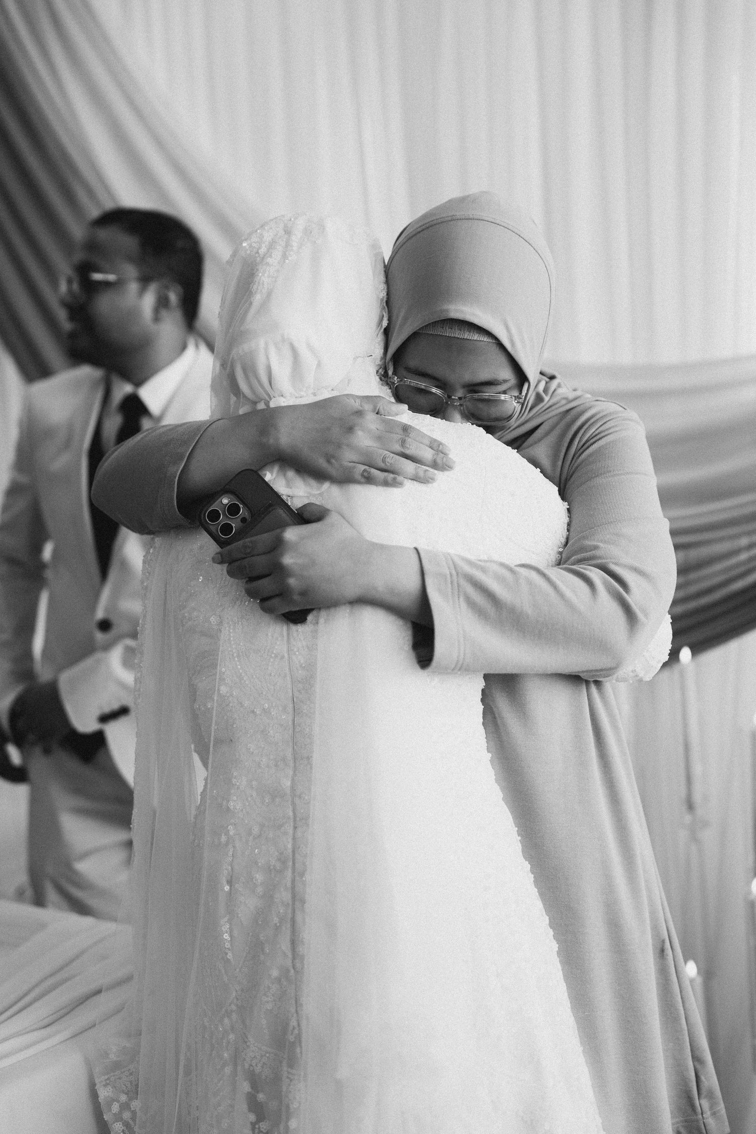 Emotional black and white photo of two women in hijabs sharing a heartfelt hug at a wedding.