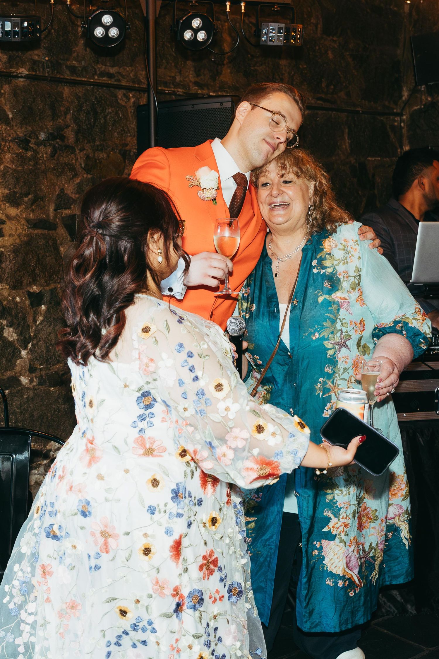 Groom in orange suit celebrates with guests in floral outfits during lively indoor wedding reception.