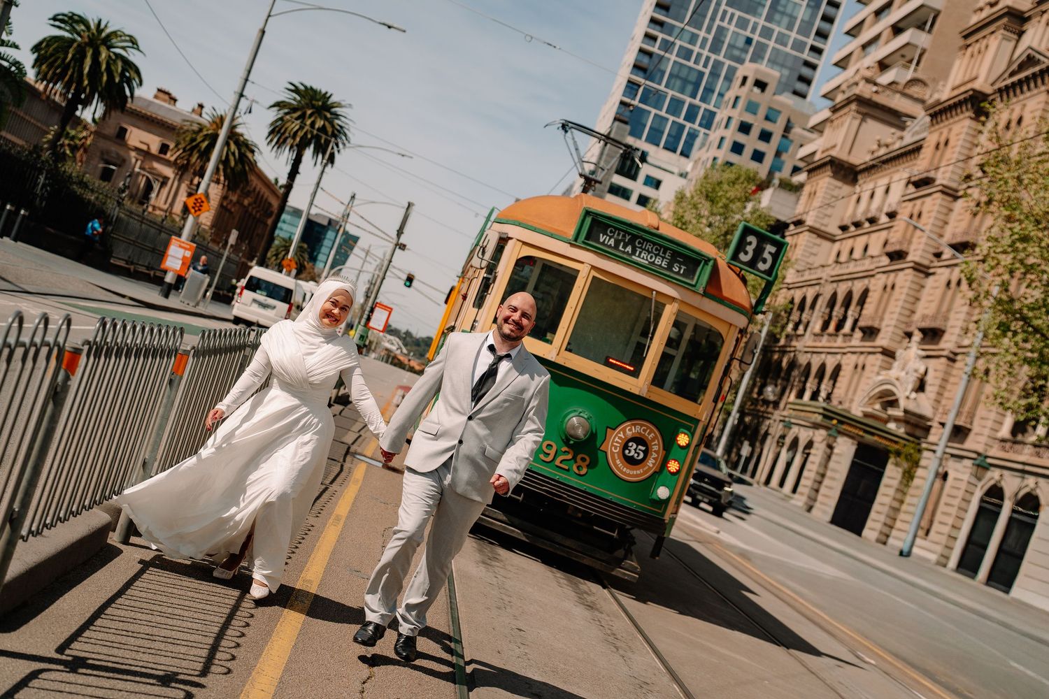 A bride and groom hold hands on a Melbourne street with a classic green and gold tram passing behind them.