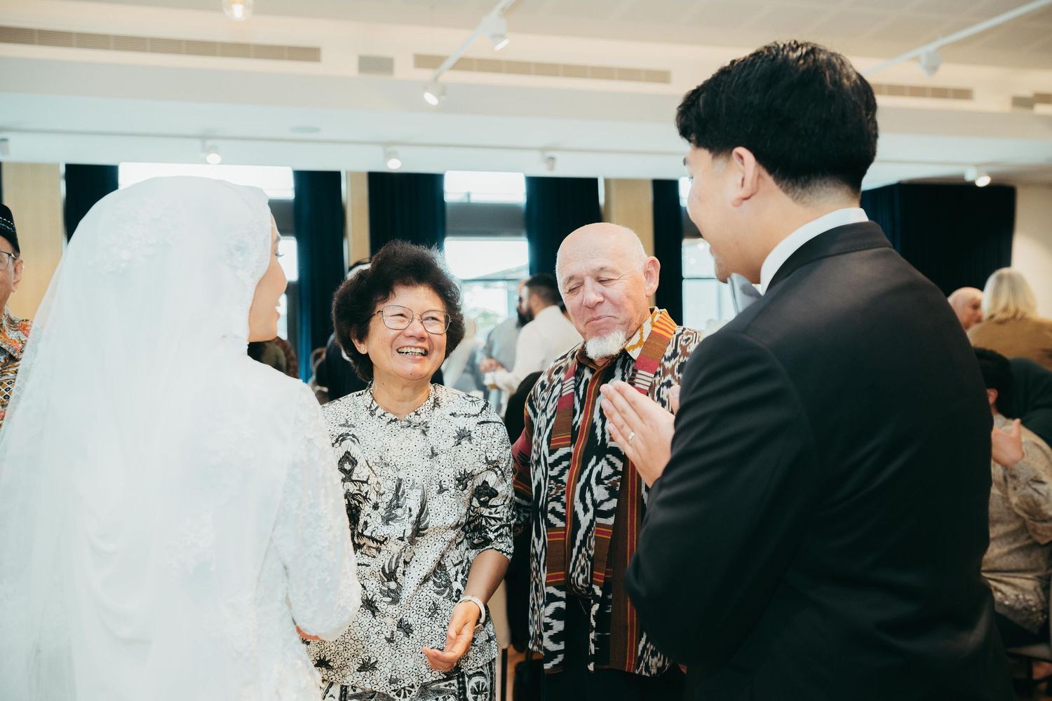 Muslim bride in white hijab and groom in suit greet smiling elderly guests at wedding reception.