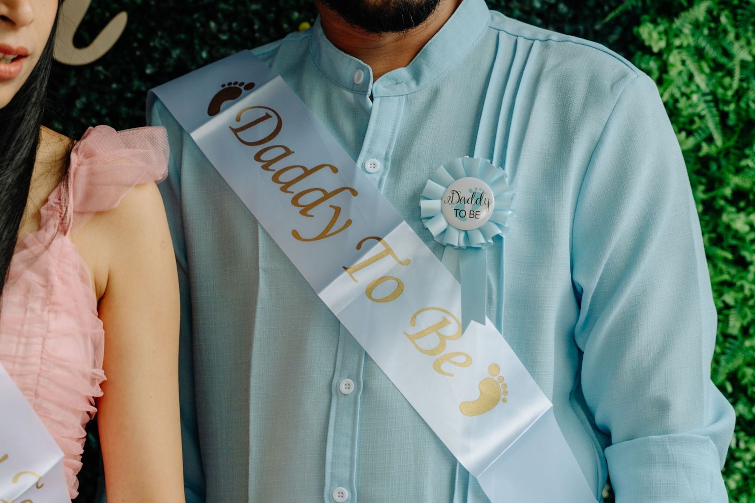 A couple in coordinated pastel outfits stands close together in front of lush green foliage.