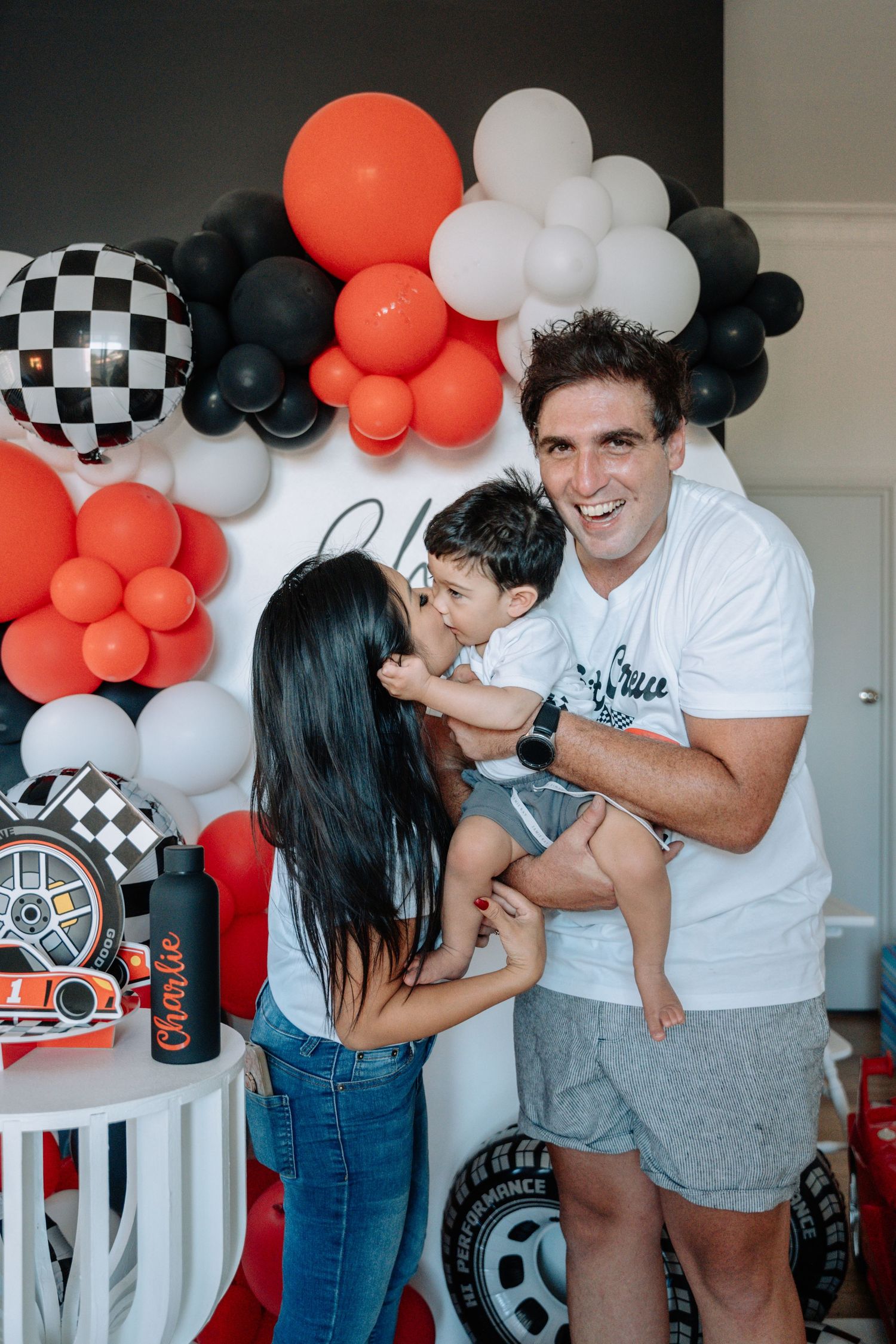 A family shares playful moments against a backdrop of orange and white balloons with racing-themed party decorations.