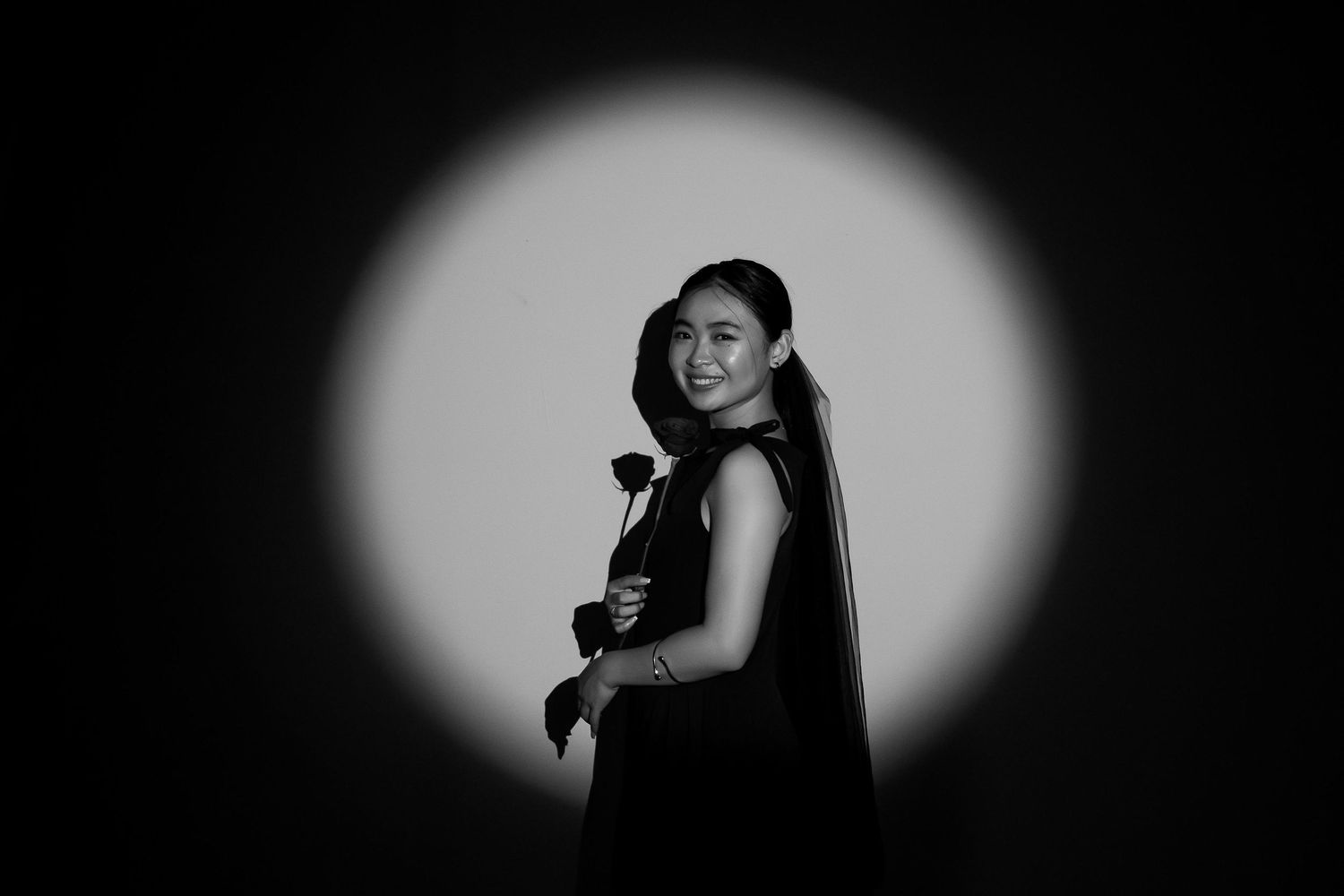 Black and white portrait of a smiling woman holding a rose, illuminated by a circular spotlight.