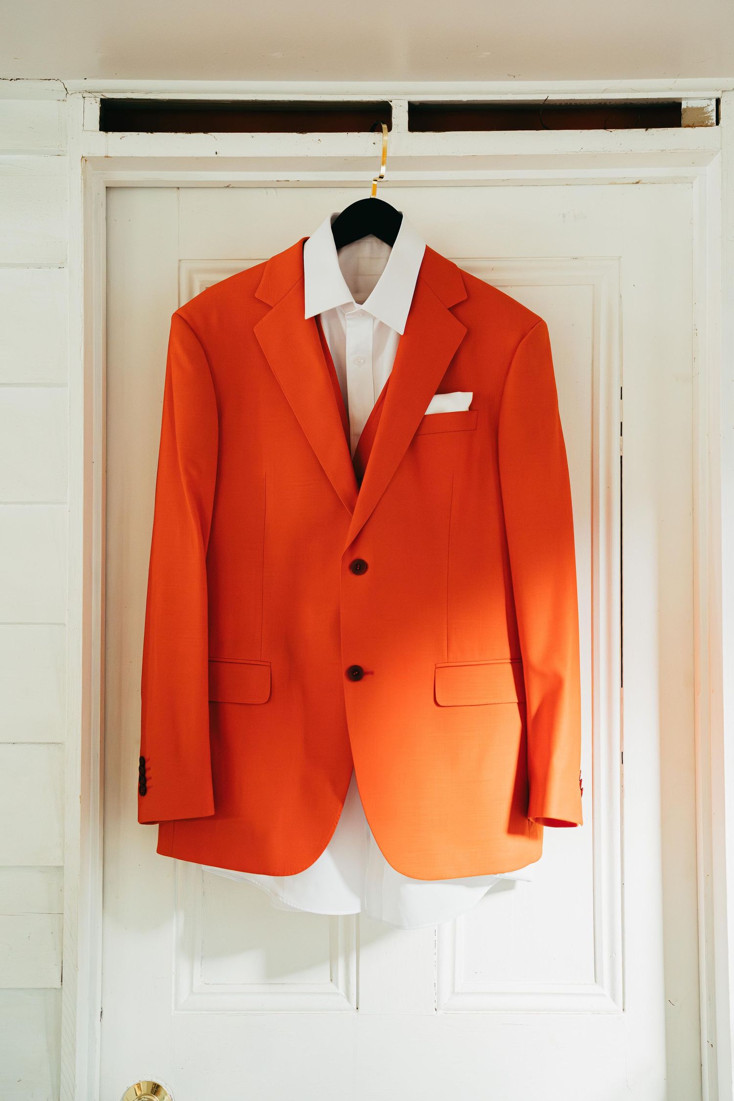 Vibrant orange wedding suit jacket with white dress shirt hanging on white door for groom preparation.