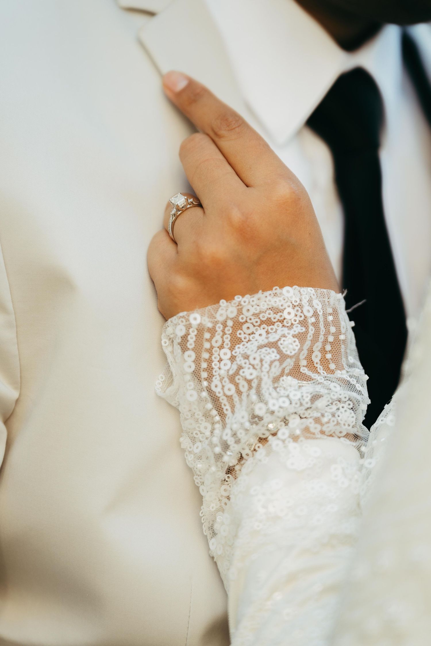 Close-up of bride's beaded lace sleeve and diamond engagement ring resting on groom's white suit jacket.