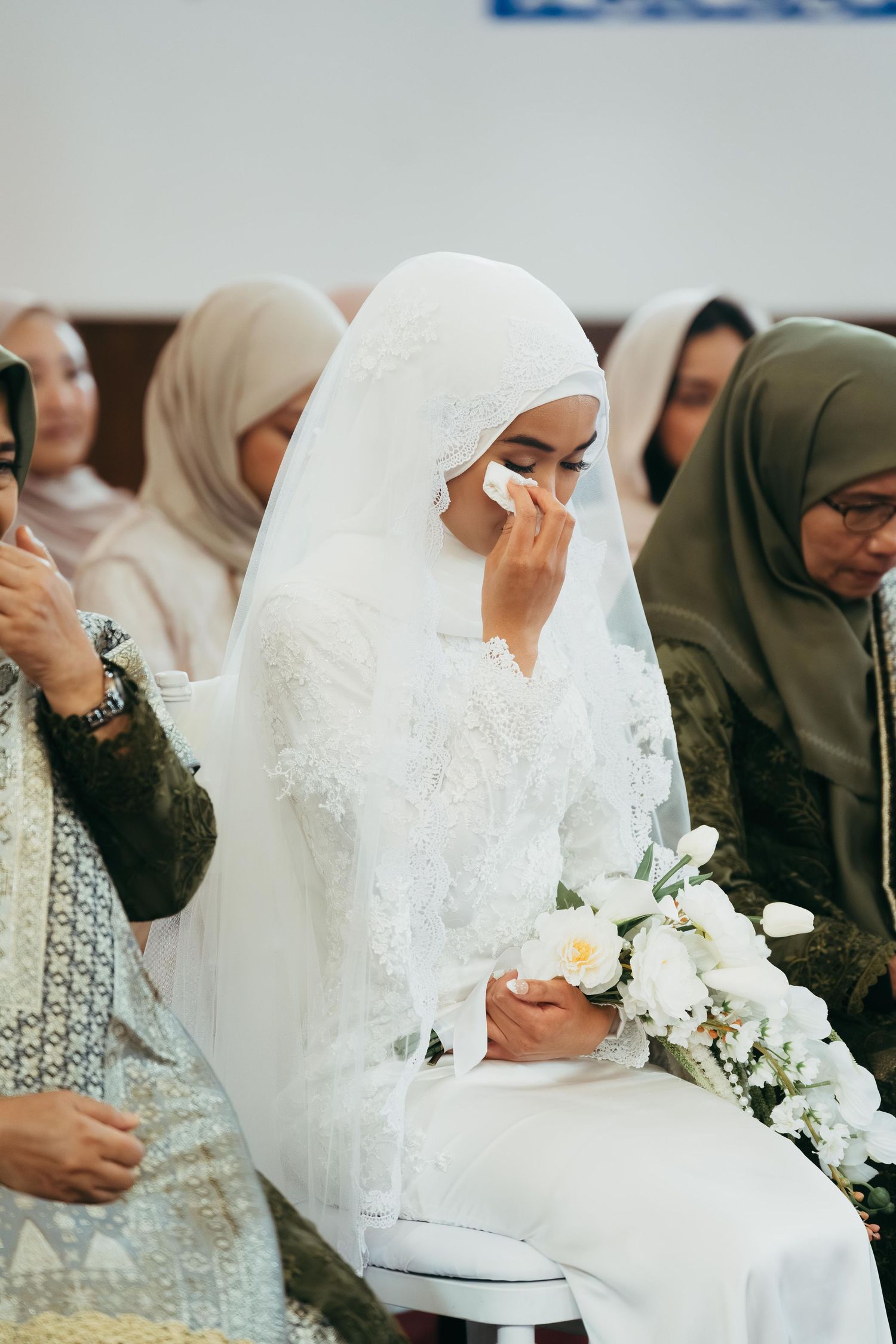 Muslim bride in white hijab and lace dress wiping tears at wedding ceremony.