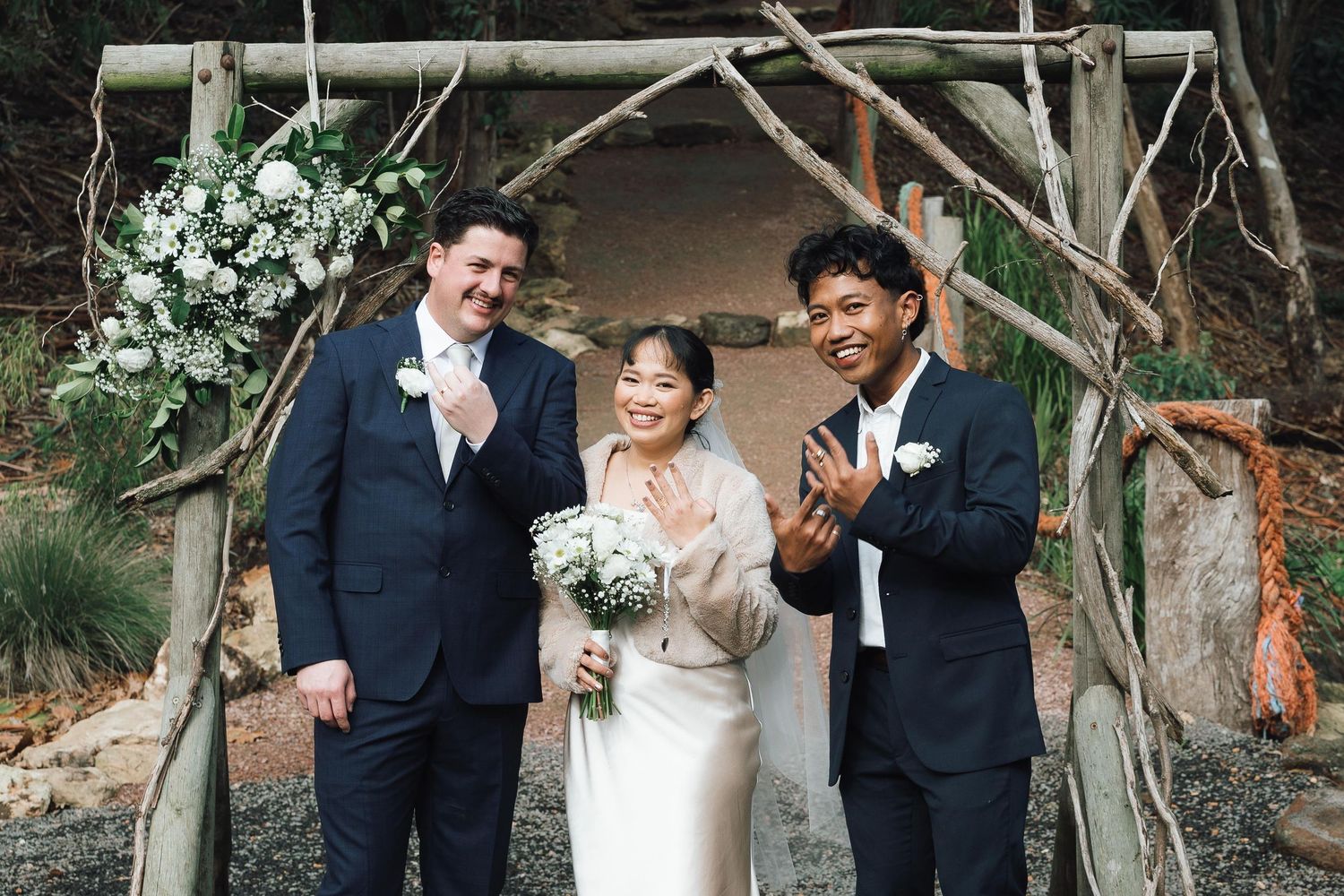 Bride and groom celebrate with officiant under rustic wooden arch adorned with white flowers at outdoor wedding ceremony.