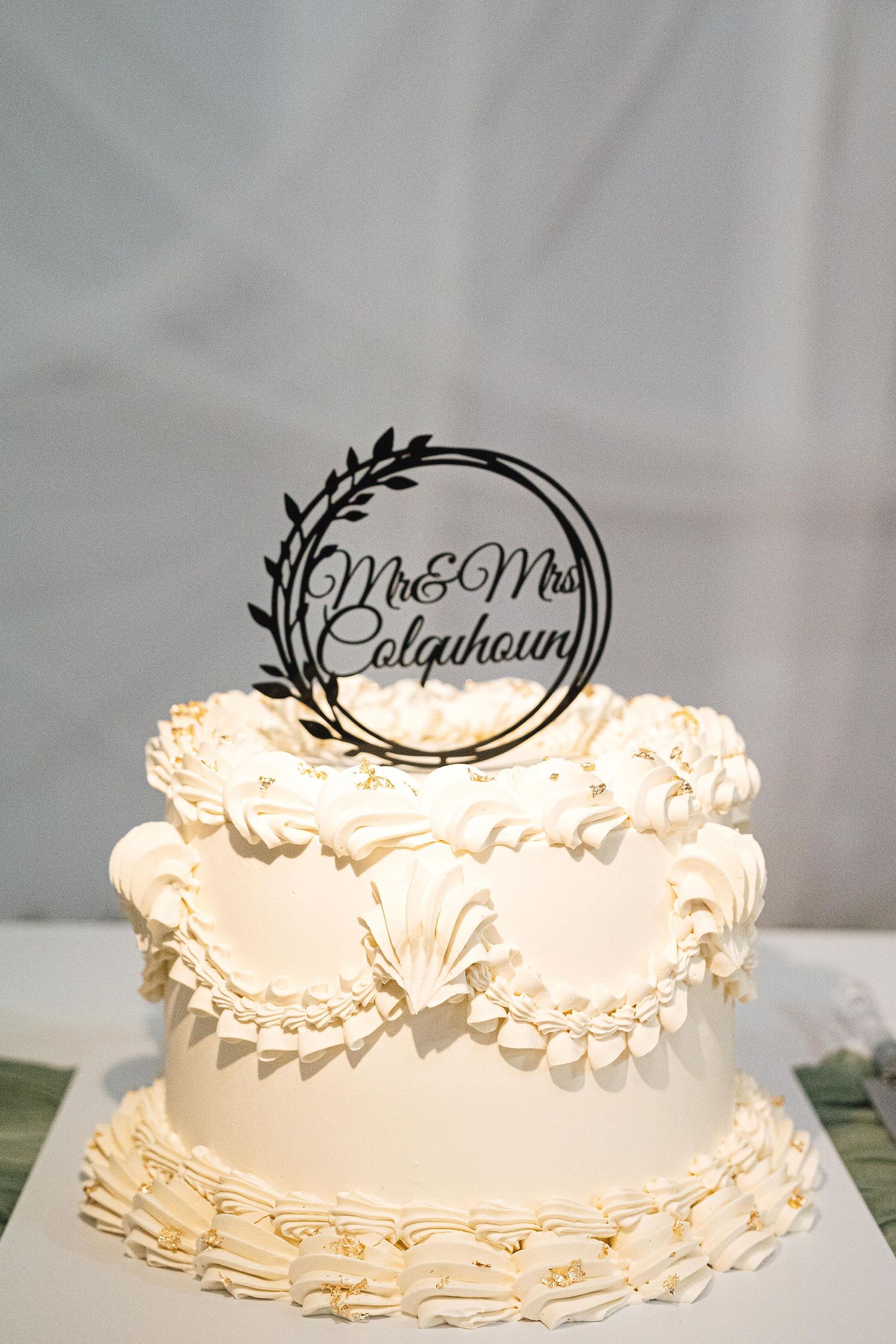 Elegant two-tier white wedding cake with ruffled frosting and Mr & Mrs Colquhoun cake topper with wreath design.