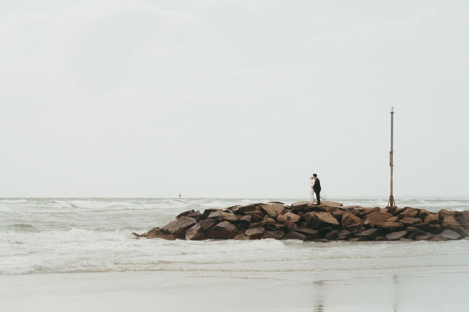Lone person standing on rocky ocean jetty under overcast sky near breaking waves.