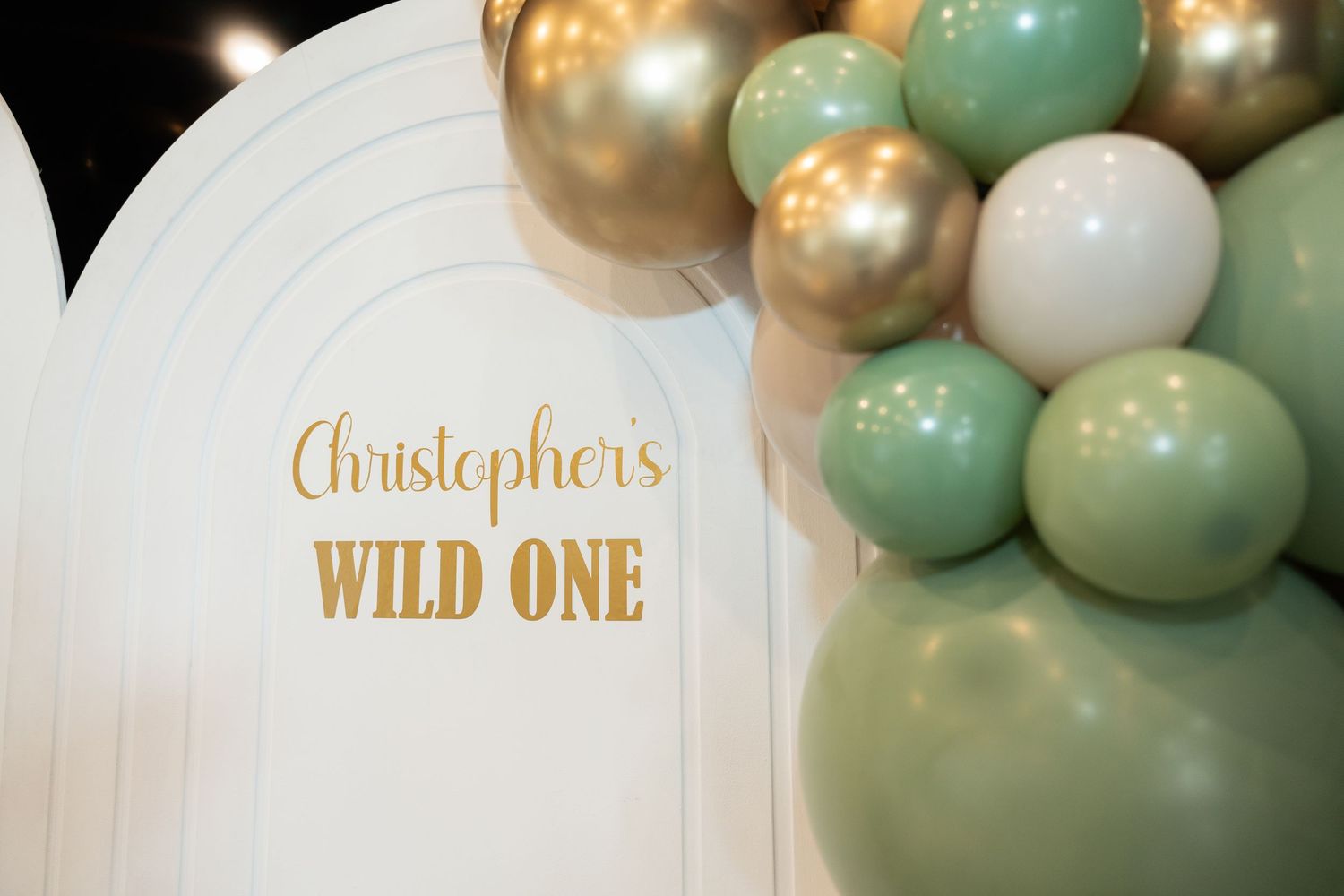 White decorative panel with gold text reading Christopher's Wild One adorned with mint green and gold balloons.