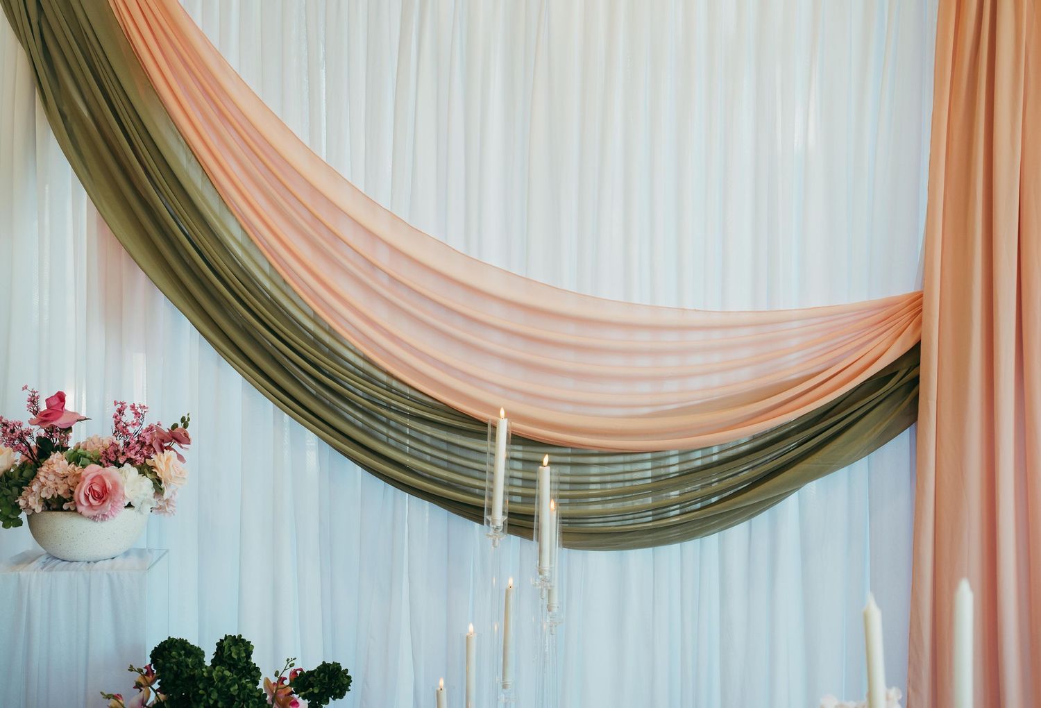 Elegant wedding backdrop with draped olive green and blush pink fabric swags, candles, and floral arrangements.