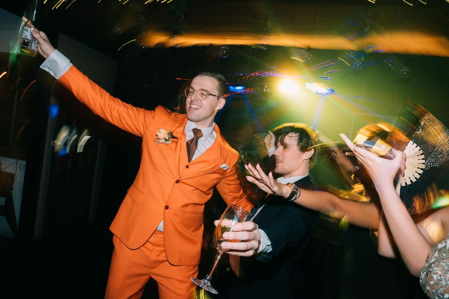 Groom in orange suit celebrating at wedding reception party with sparklers.