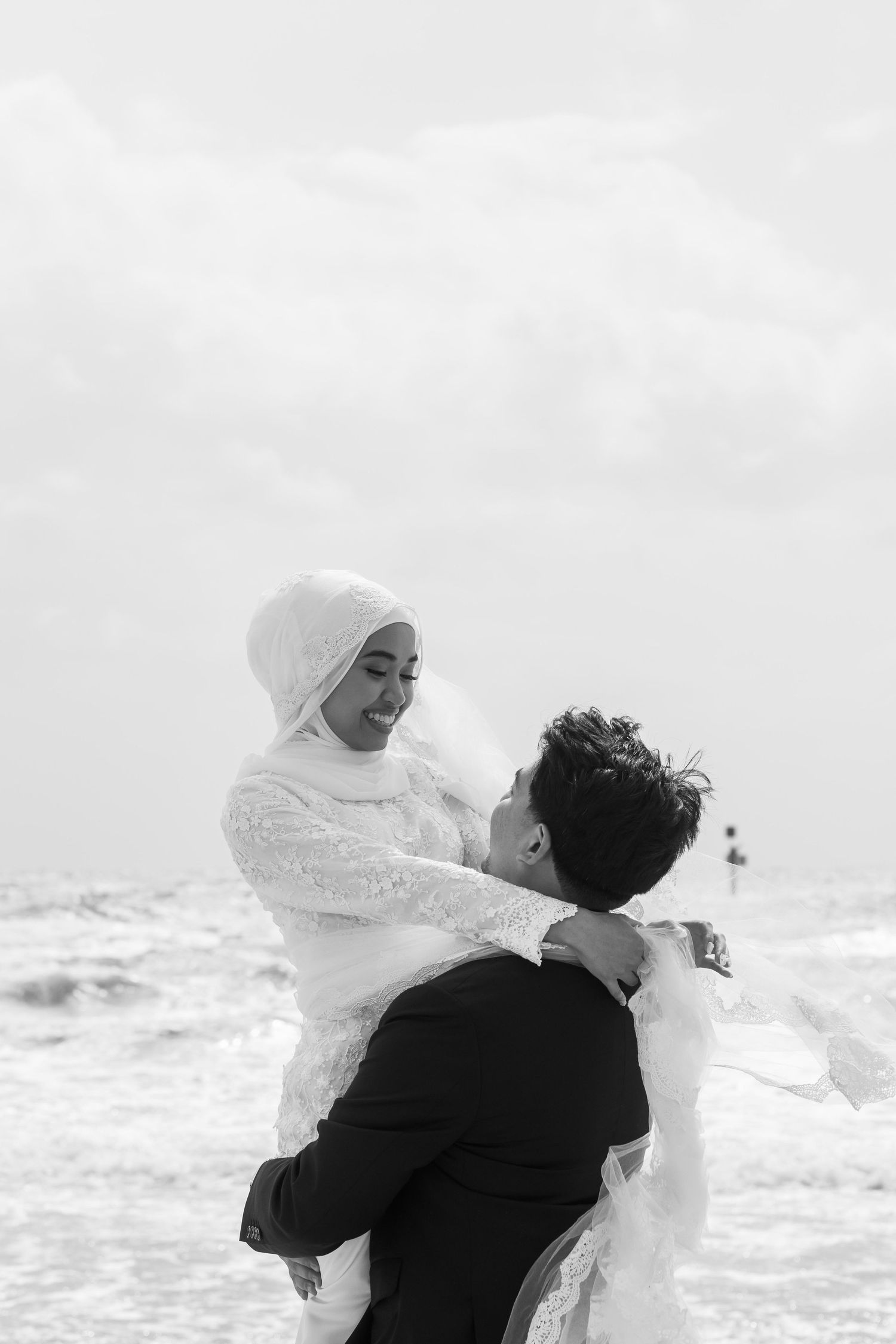 Black and white photo of a groom lifting his bride in a hijab by the ocean on their wedding day.