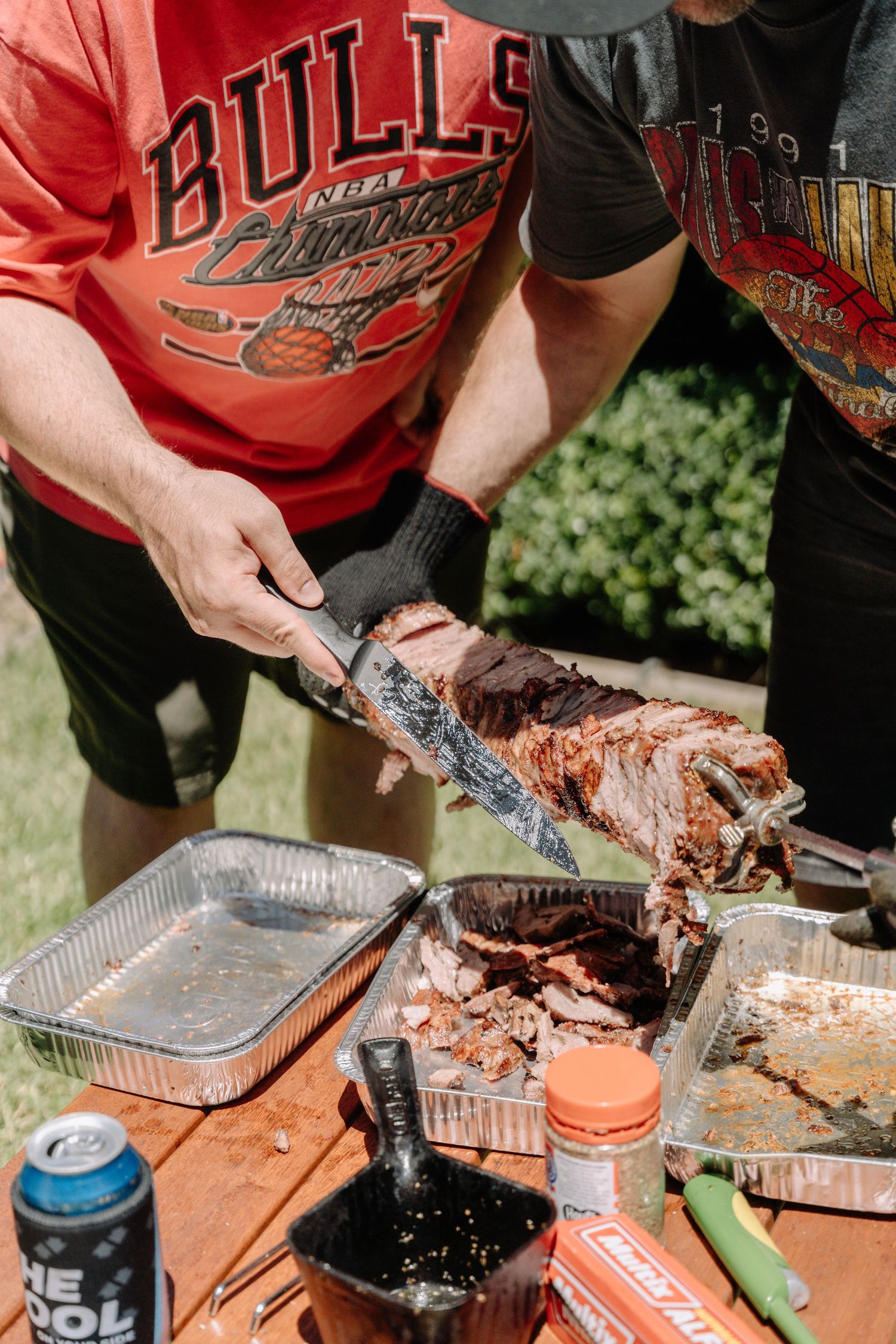 Close-up of hands cutting barbecued meat on a wooden table with condiments and drinks nearby.