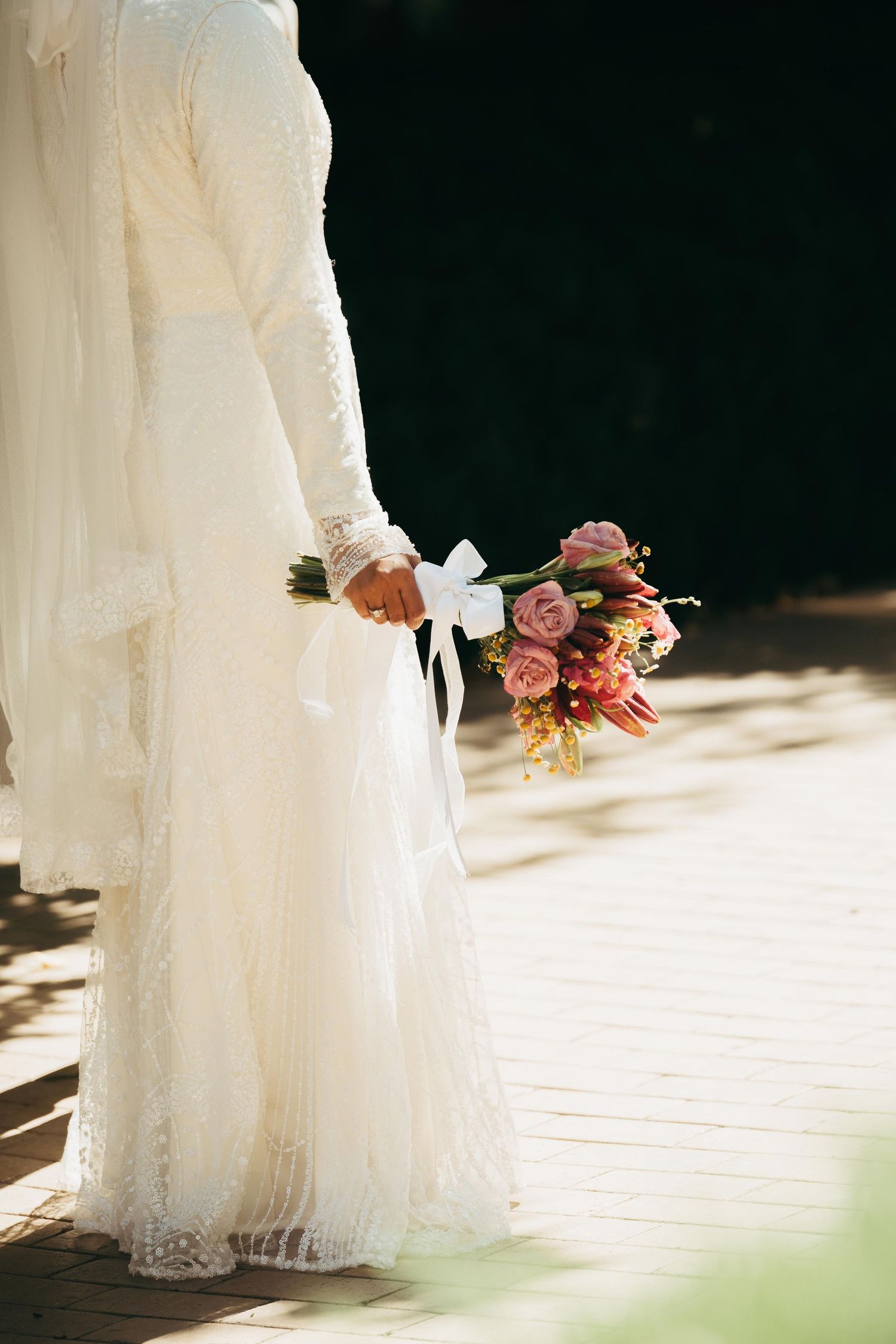 Bride in white lace long-sleeve wedding dress holding a pink and red bridal bouquet outdoors.