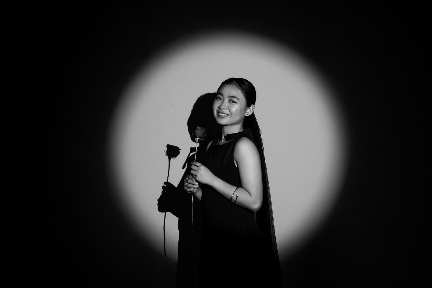 Black and white portrait of a smiling woman holding a rose in a dramatic spotlight on dark background.