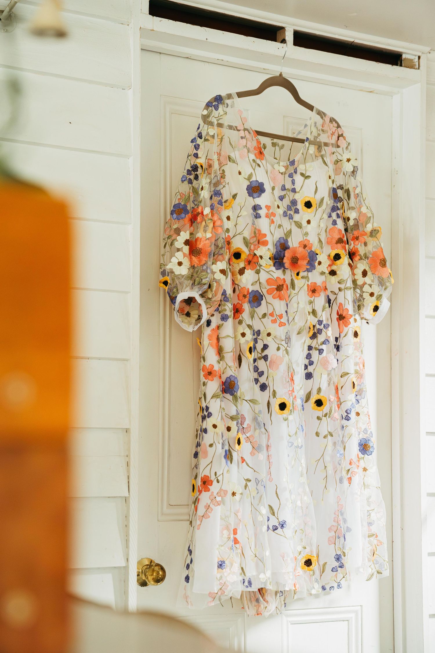 A floral embroidered white wedding dress hanging on a wooden door in a bright white room.