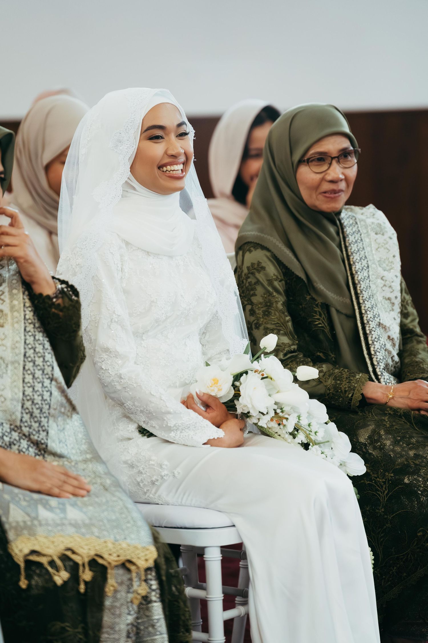 Radiant Muslim bride in white lace wedding dress holding bouquet smiling during ceremony.