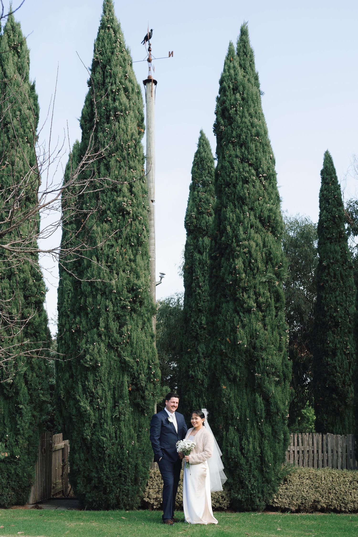 Bride and groom stand together on green lawn surrounded by tall cypress trees on their wedding day.