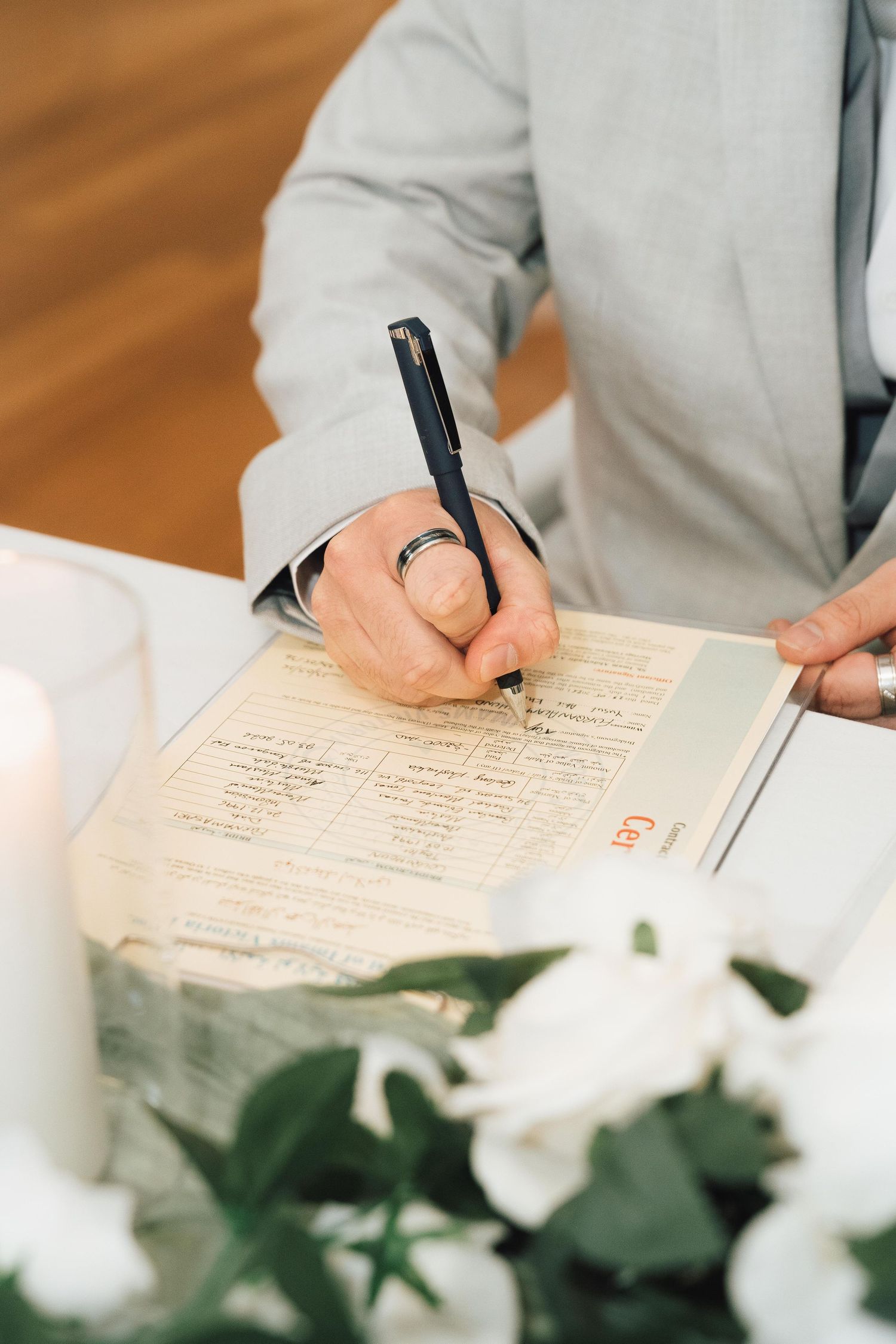 Person in gray suit signing wedding registry document with black pen, wearing a silver ring, with white flowers in foreground.