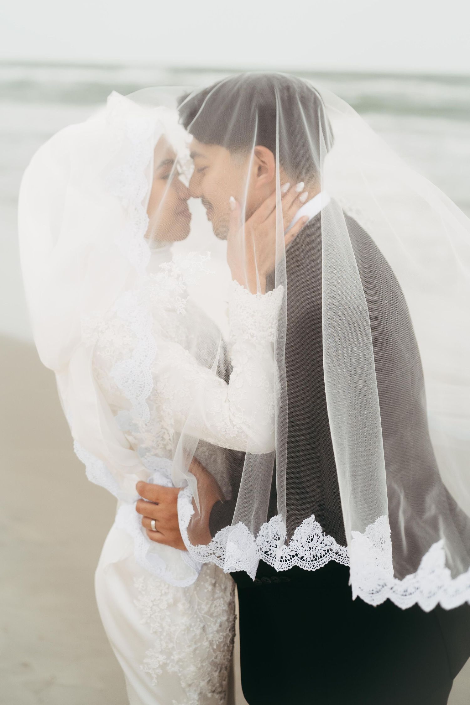 Muslim bride and groom share a romantic moment under a lace-trimmed veil by the beach.