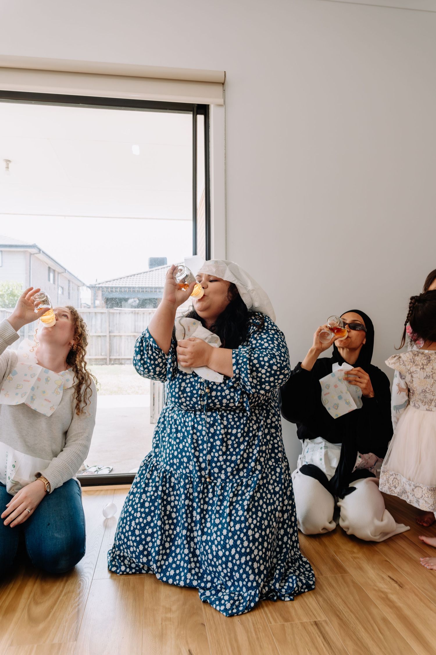 People in patterned dresses and white outfits strike theatrical poses on a wooden floor in a bright room.