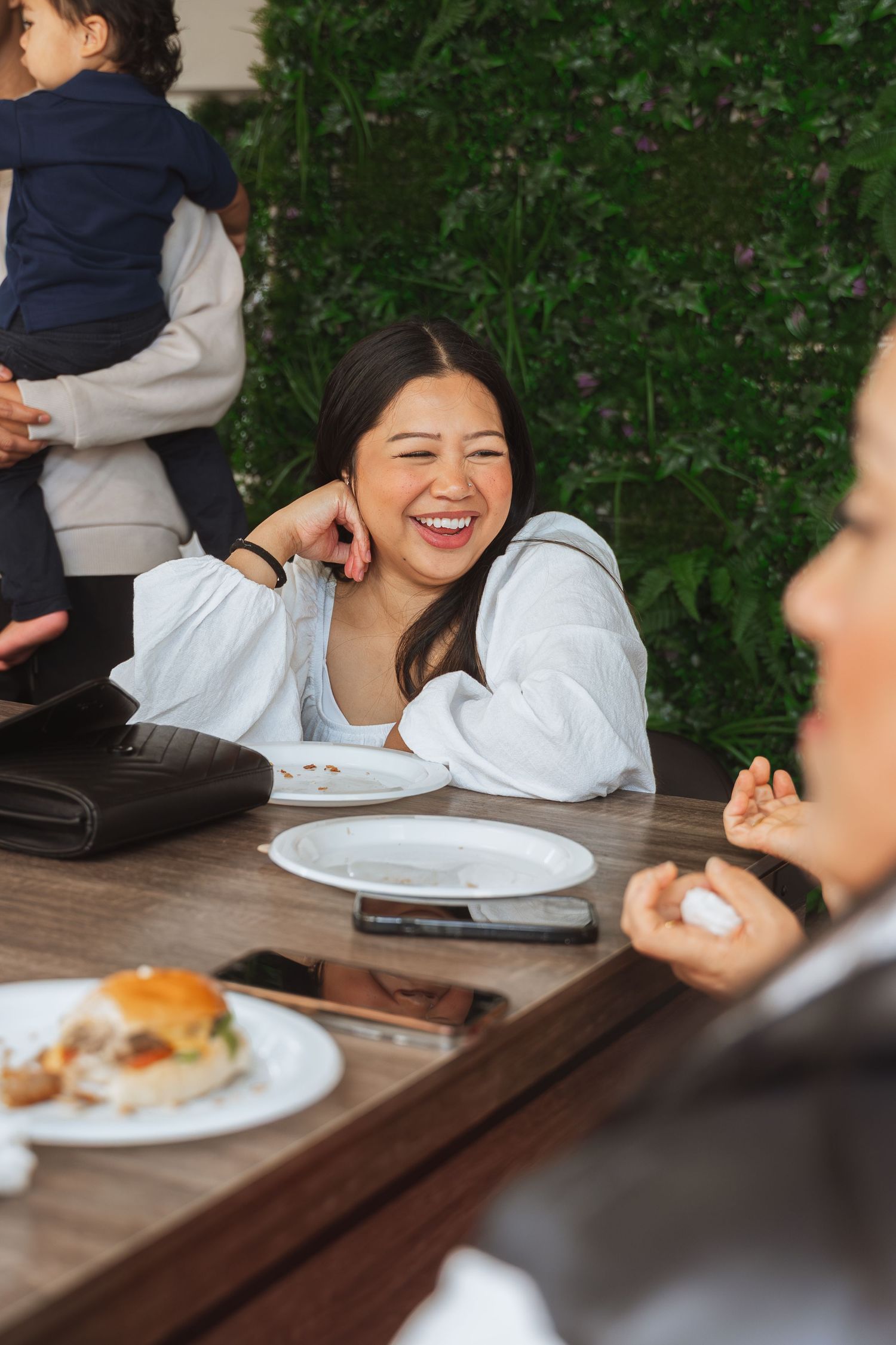 People enjoying a casual outdoor meal at a wooden picnic table with plates of food and laughter.